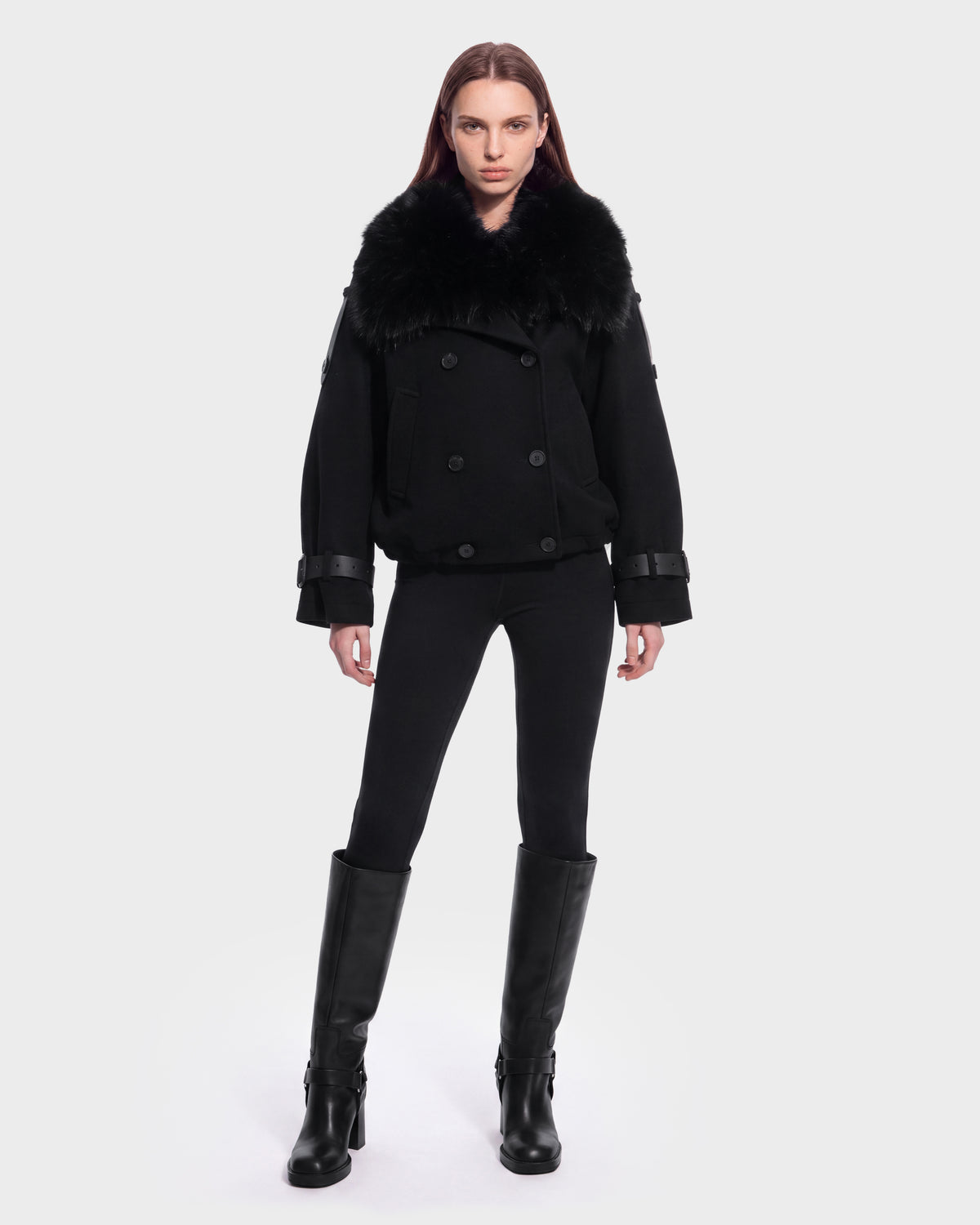 A person stands facing forward against a plain white background, wearing the Rudsak MAEMI - 8126985 Black double-breasted coat with a large removable faux fur collar, paired with black leggings and knee-high black boots.