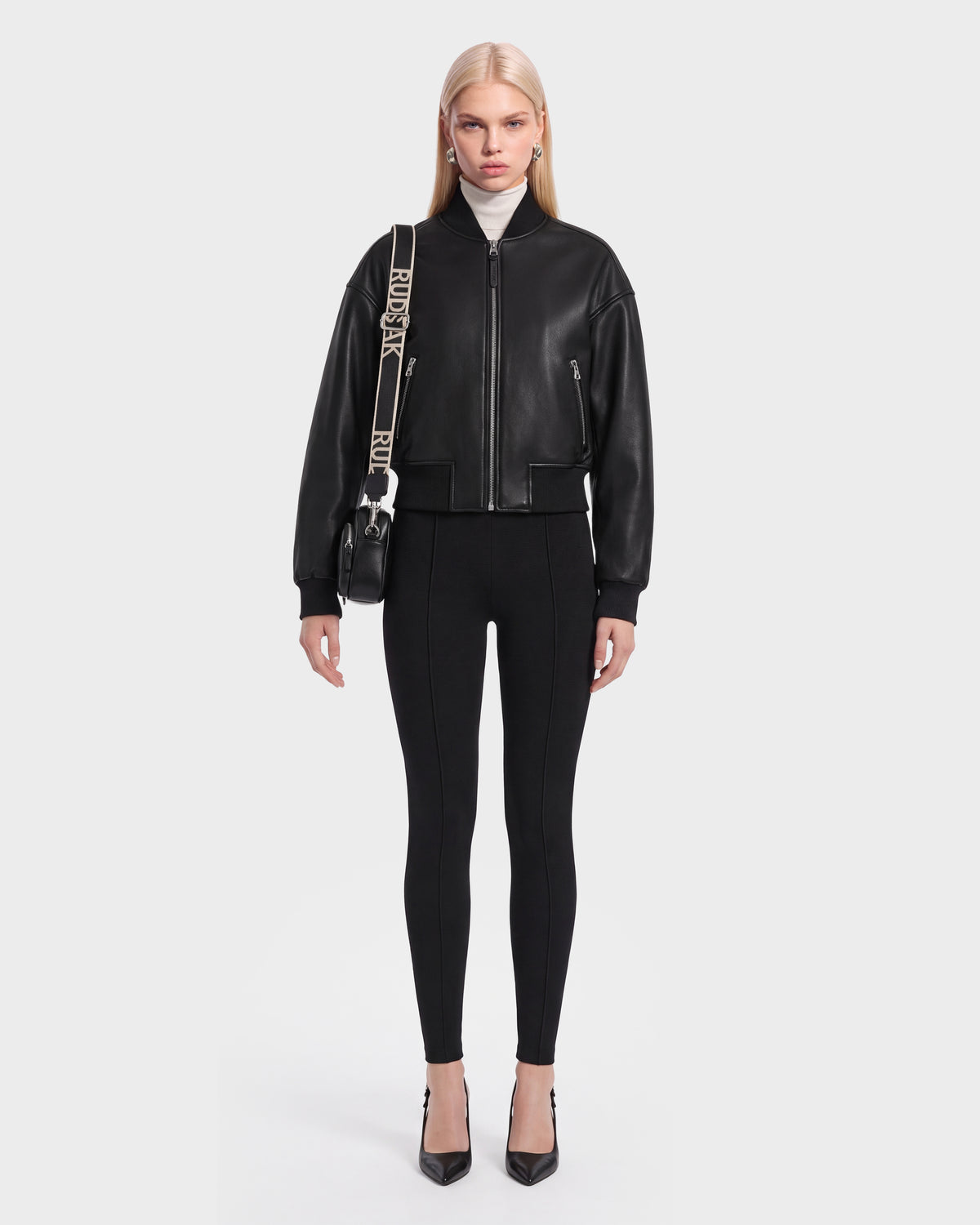 A woman stands facing forward, wearing the Rudsak EMERSON - 8125041 Black cropped silhouette leather bomber jacket styled with black leggings, black heels, and a small crossbody bag with a bold strap. She has straight blonde hair and a neutral expression against a plain white background, showcasing this versatile wardrobe staple.