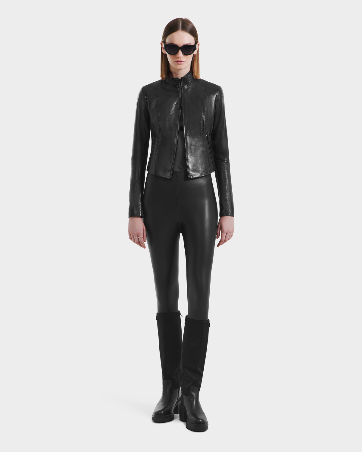 A woman wearing large sunglasses, a fitted Rudsak ABIGAIL N - 8125970 Black leather jacket, black leggings, and black knee-high boots stands against a plain white background.