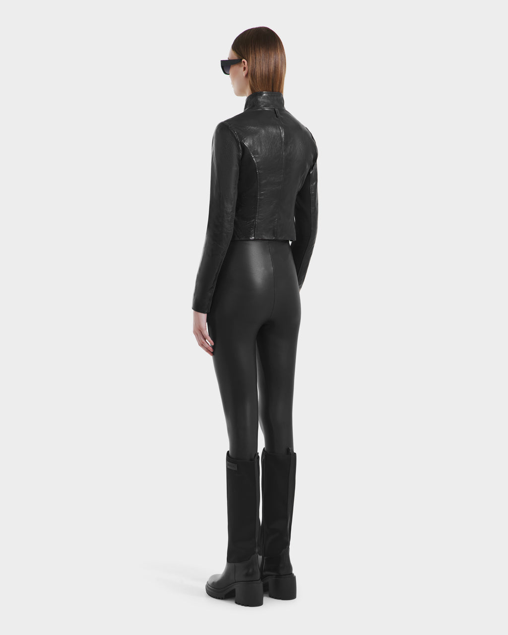 A person faces away, wearing the ABIGAIL N - 8125970 Black leather racer jacket by Rudsak, paired with black leggings, boots, and sunglasses. The image is set against a plain white background.