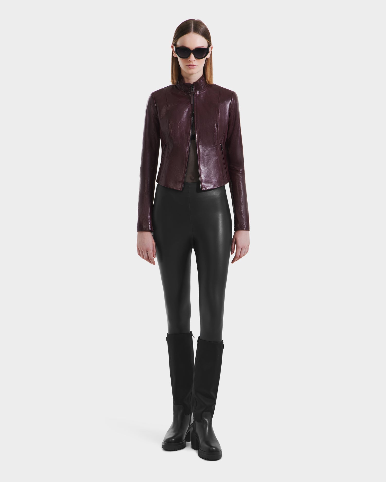 A person wearing black sunglasses, the Rudsak ABIGAIL - 8124560 Bordeaux leather jacket, black leggings, and black knee-high boots stands against a plain white background.
