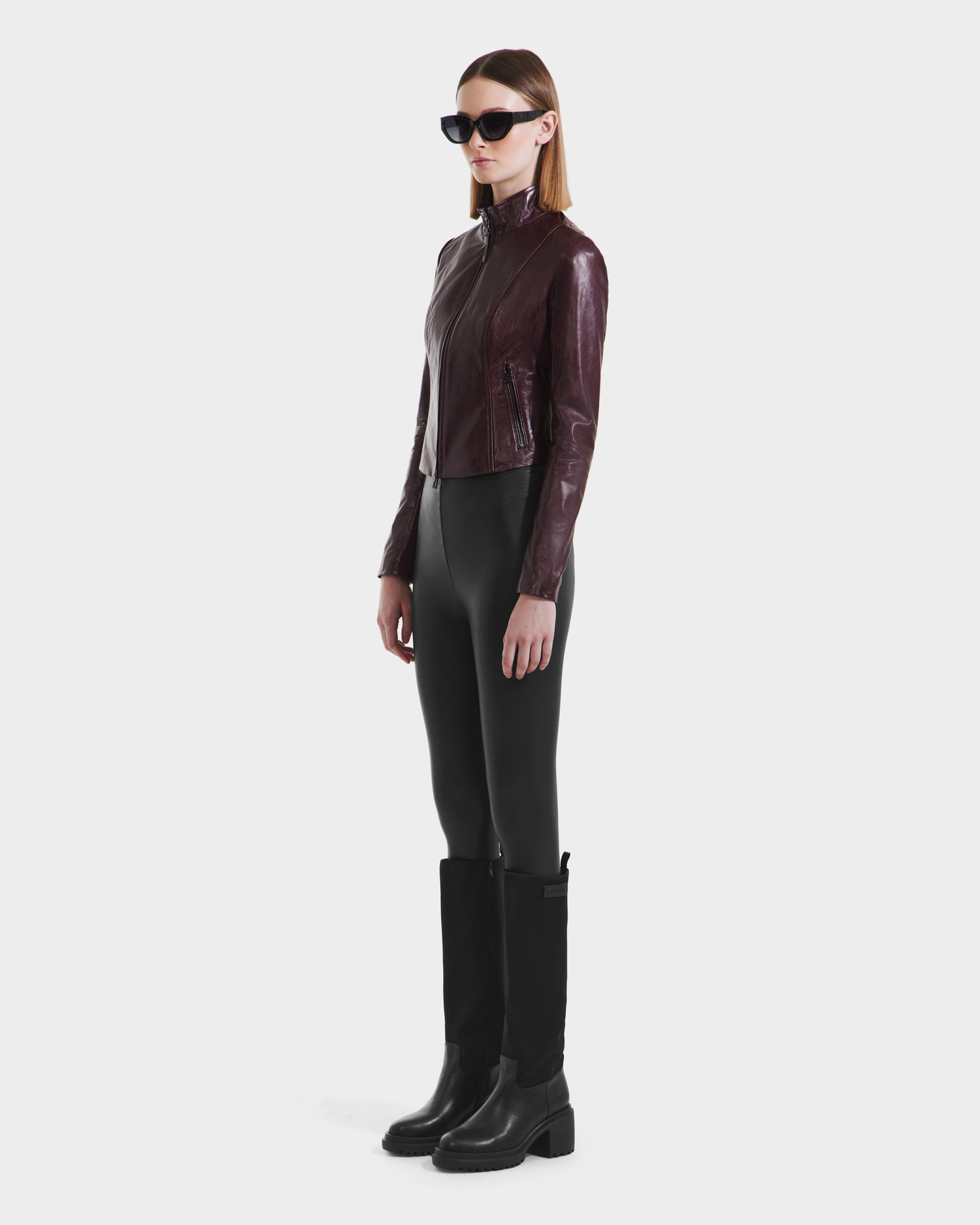 A woman in black sunglasses, the ABIGAIL - 8124560 Bordeaux leather racer jacket by Rudsak, black leggings, and knee-high black boots stands against a plain white background.
