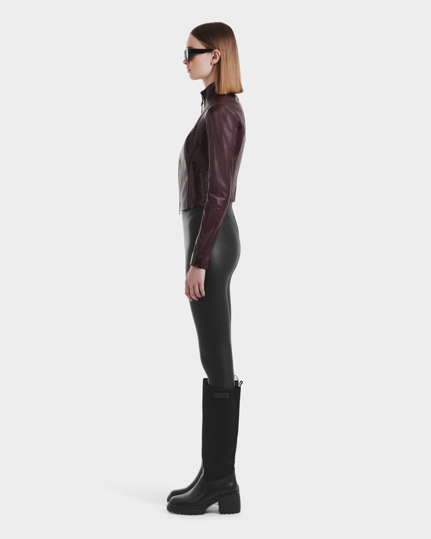 A woman with straight, shoulder-length brown hair stands in profile against a white background, wearing the Rudsak ABIGAIL - 8124560 Bordeaux leather racer jacket, paired with black leggings, tall black boots, and sunglasses.