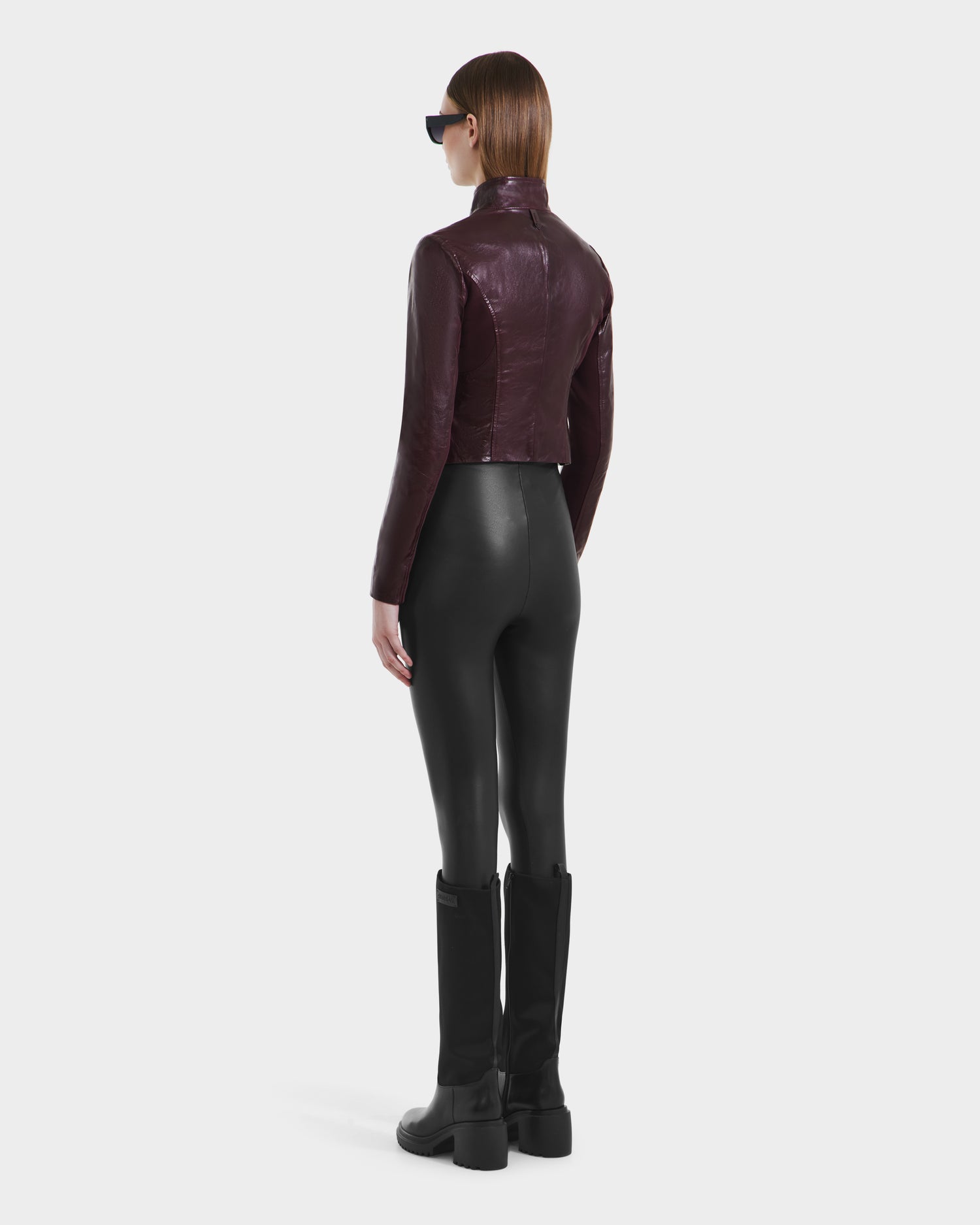 A person with straight brown hair, black sunglasses, and the Rudsak ABIGAIL - 8124560 Bordeaux womens leather racer jacket, paired with fitted black pants and boots, stands facing away against a plain white background.