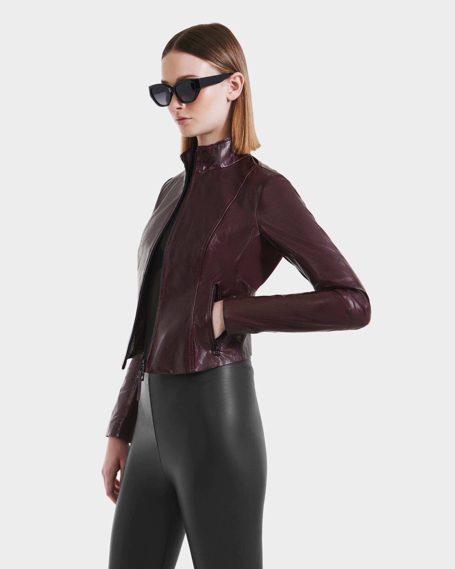 A woman in black sunglasses, a Rudsak ABIGAIL - 8124560 Bordeaux leather jacket, and high-waisted black leggings stands against a plain white background.