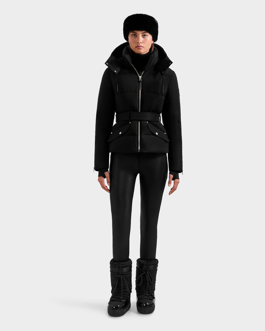 A person stands facing forward in a black Rudsak ADELINE H - 8125587 women's belted down puffer jacket, paired with black pants, black winter boots, and a black fur hat, set against a plain light gray background.