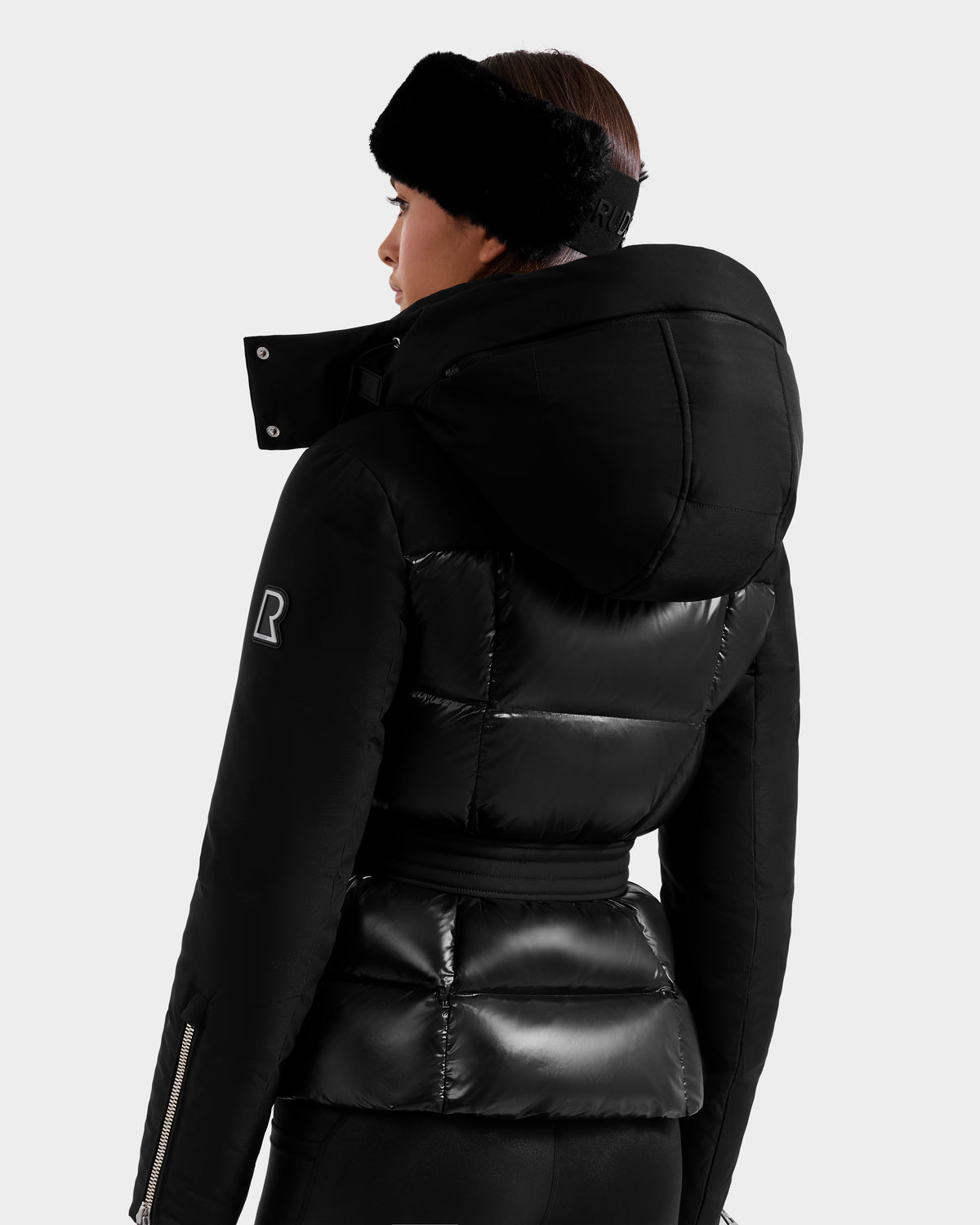 A woman, shown from the back against a white background, wears the Rudsak ADELINE H - 8125587 Black jacket with a hood, paired with a black headband and pants. This sleek women’s down puffer is perfect for winter.