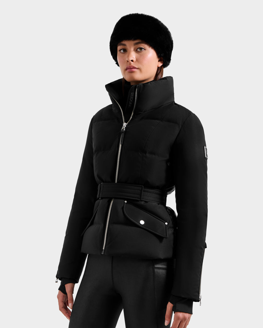 A woman in the Rudsak ADELINE H - 8125587 Black jacket, with a belt, a black faux fur hat, and black leather pants, stands against a plain light gray background, looking straight ahead with a neutral expression.