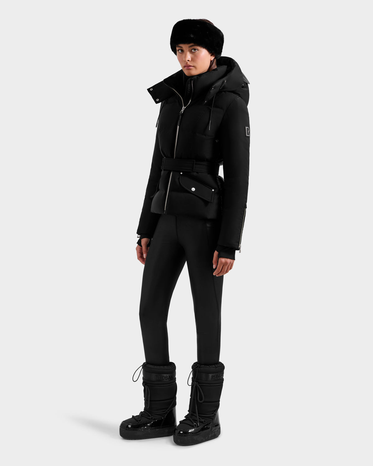 A woman wearing the Rudsak ADELINE H - 8125587 Black jacket with a belt, black pants, a black fur hat, and winter boots stands against a plain light background.