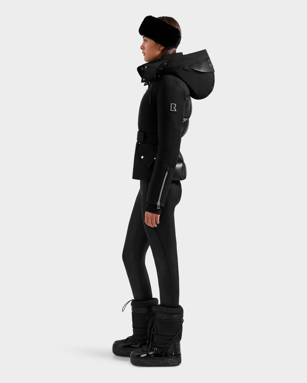 A person in profile wears the Rudsak ADELINE H - 8125587 Black jacket with a large hood, paired with black leggings, fur-lined boots, and a black headband, standing against a plain light background.