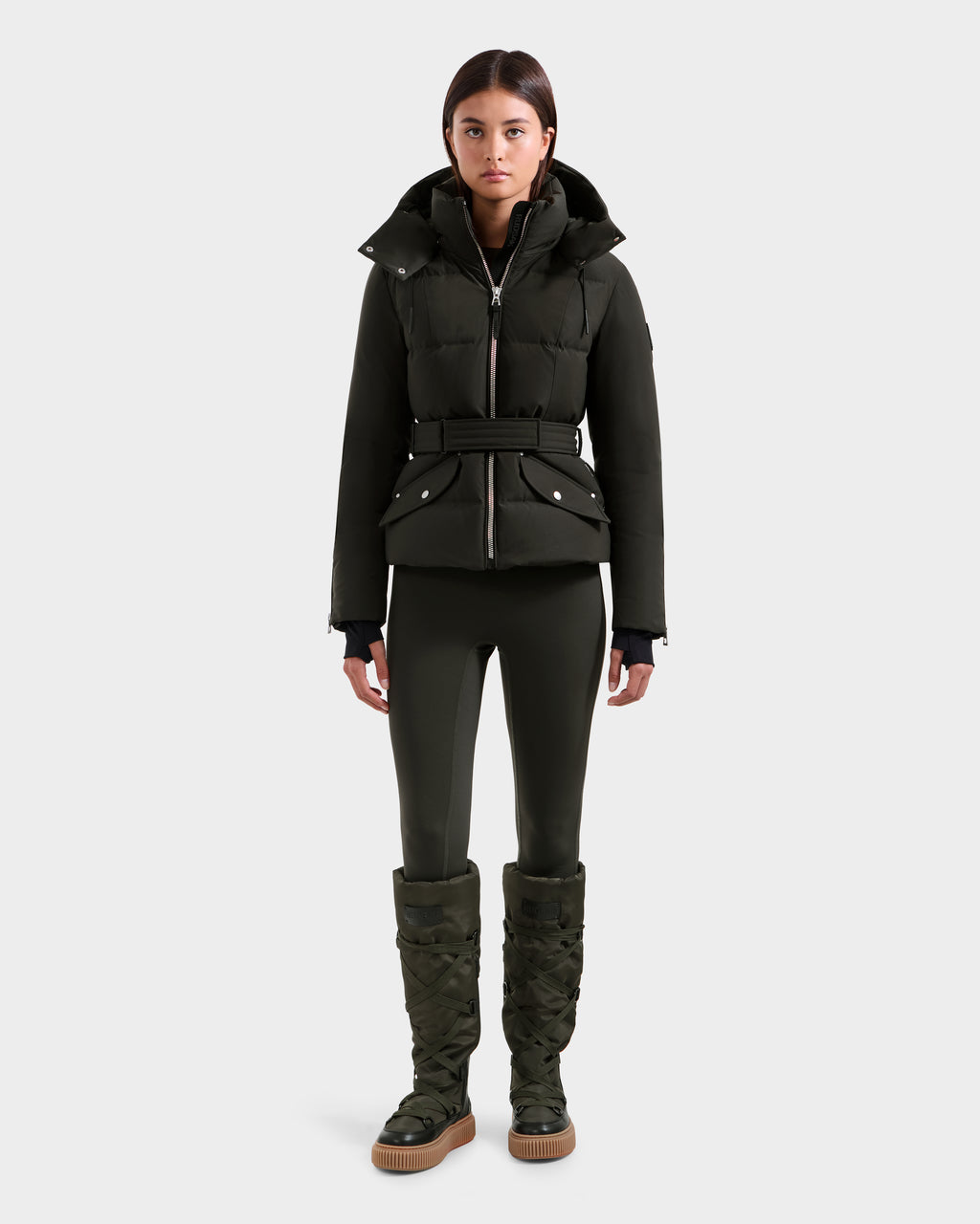 A woman stands facing forward, wearing the Rudsak ADELINE H - 8125587 down puffer jacket in Dark Olive with a belted waist, matching fitted pants, and tall quilted boots against a plain light gray background.