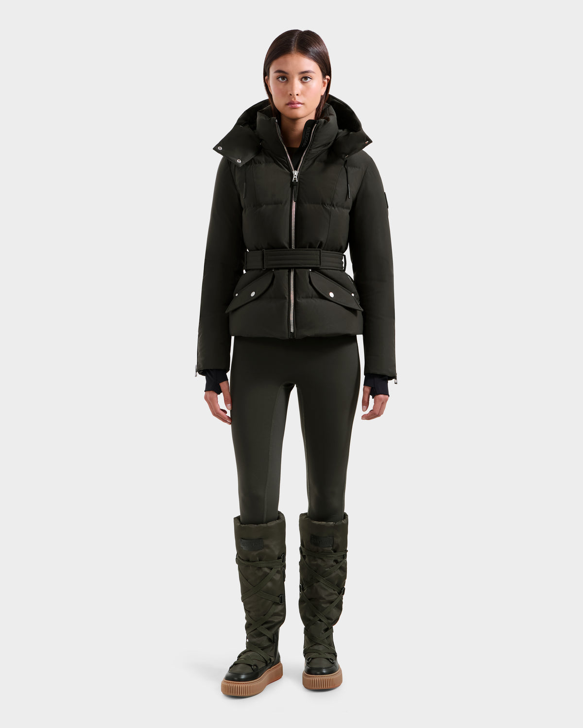 A woman stands facing forward, wearing the Rudsak ADELINE H - 8125587 down puffer jacket in Dark Olive with a belted waist, matching fitted pants, and tall quilted boots against a plain light gray background.