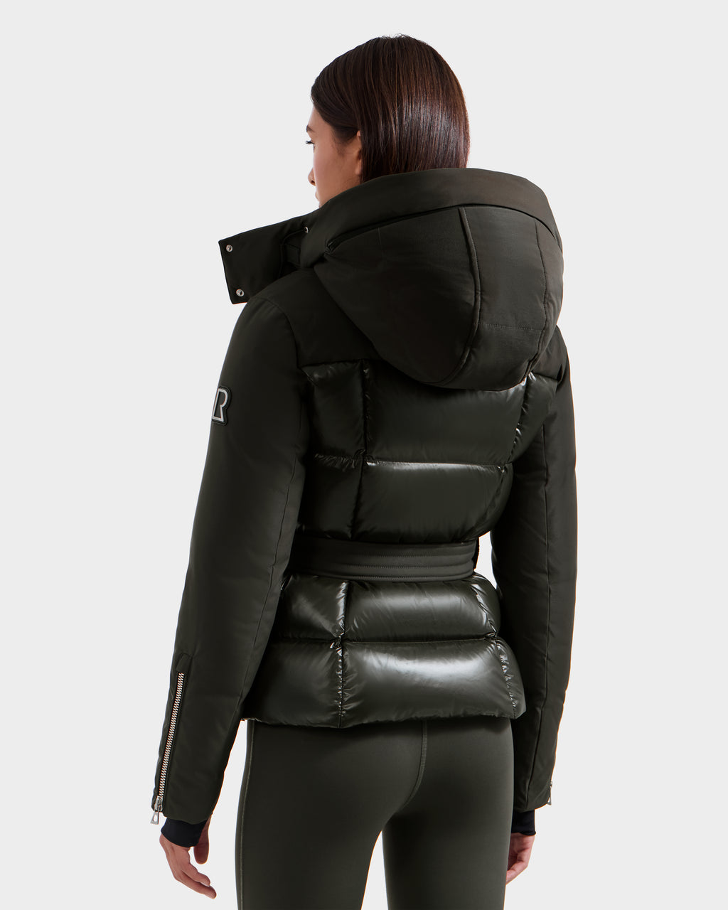 A woman is seen from behind wearing the Rudsak ADELINE H - 8125587 Dark Olive winter jacket with matching leggings, showcasing its quilted sections and fitted puffer-style design.