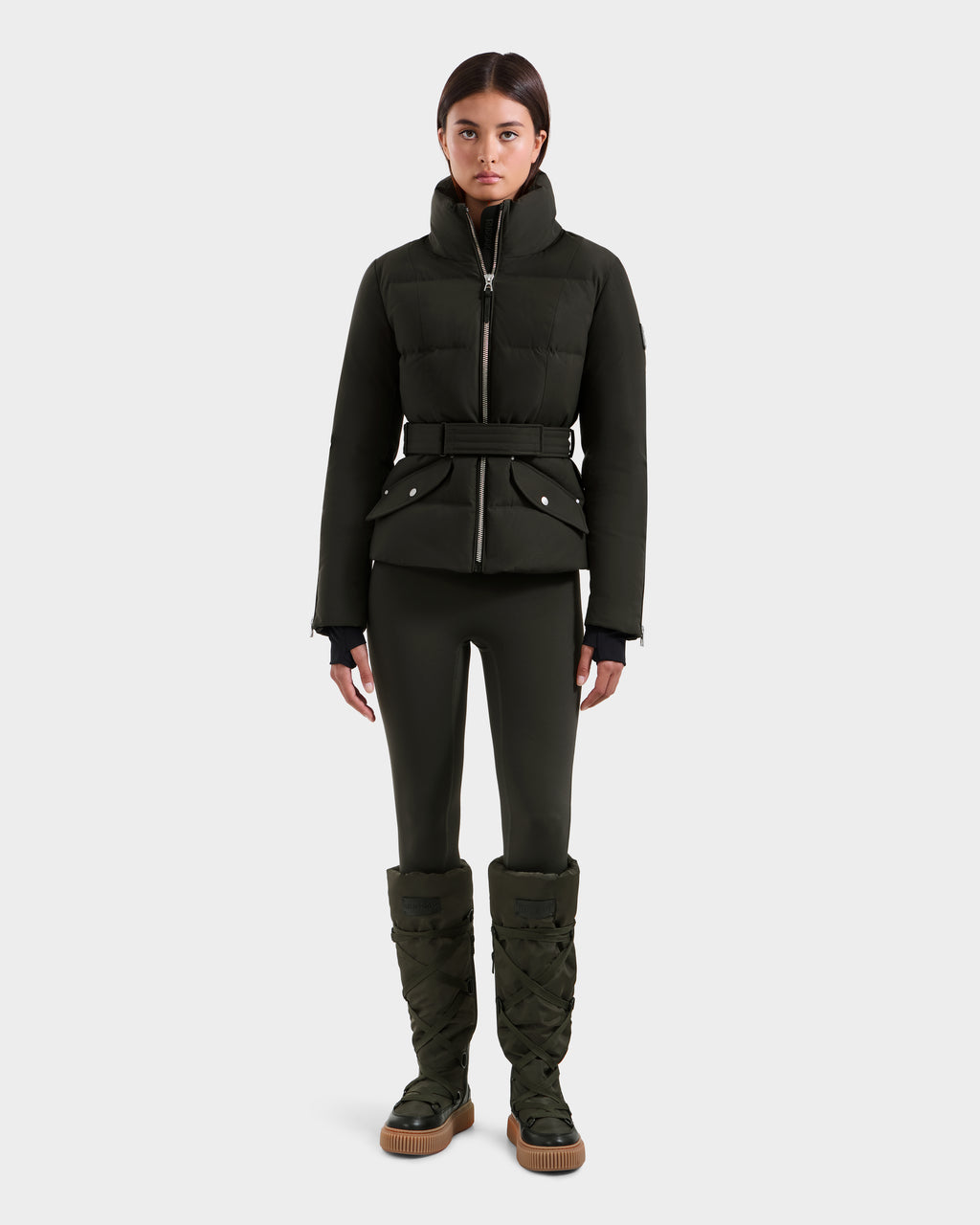 A woman faces forward wearing the Rudsak ADELINE H - 8125587 down puffer jacket in dark olive with a belt, matching fitted pants, and tall quilted boots, all against a plain white background.