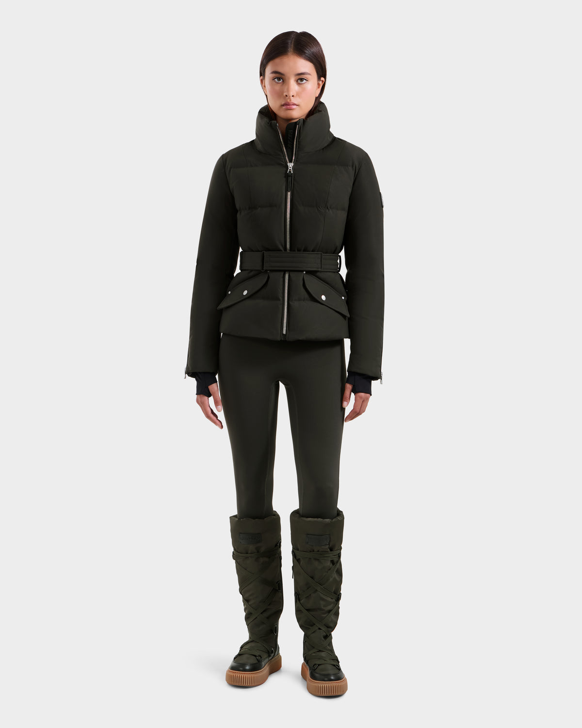 A woman faces forward wearing the Rudsak ADELINE H - 8125587 down puffer jacket in dark olive with a belt, matching fitted pants, and tall quilted boots, all against a plain white background.