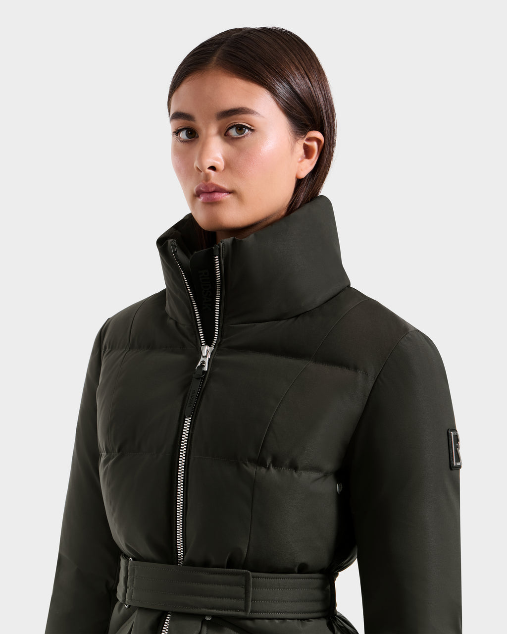 A woman with straight brown hair wears a Rudsak ADELINE H - 8125587 dark olive women’s down puffer jacket with a high collar, zipper, and matching belt, standing against a plain light gray background.