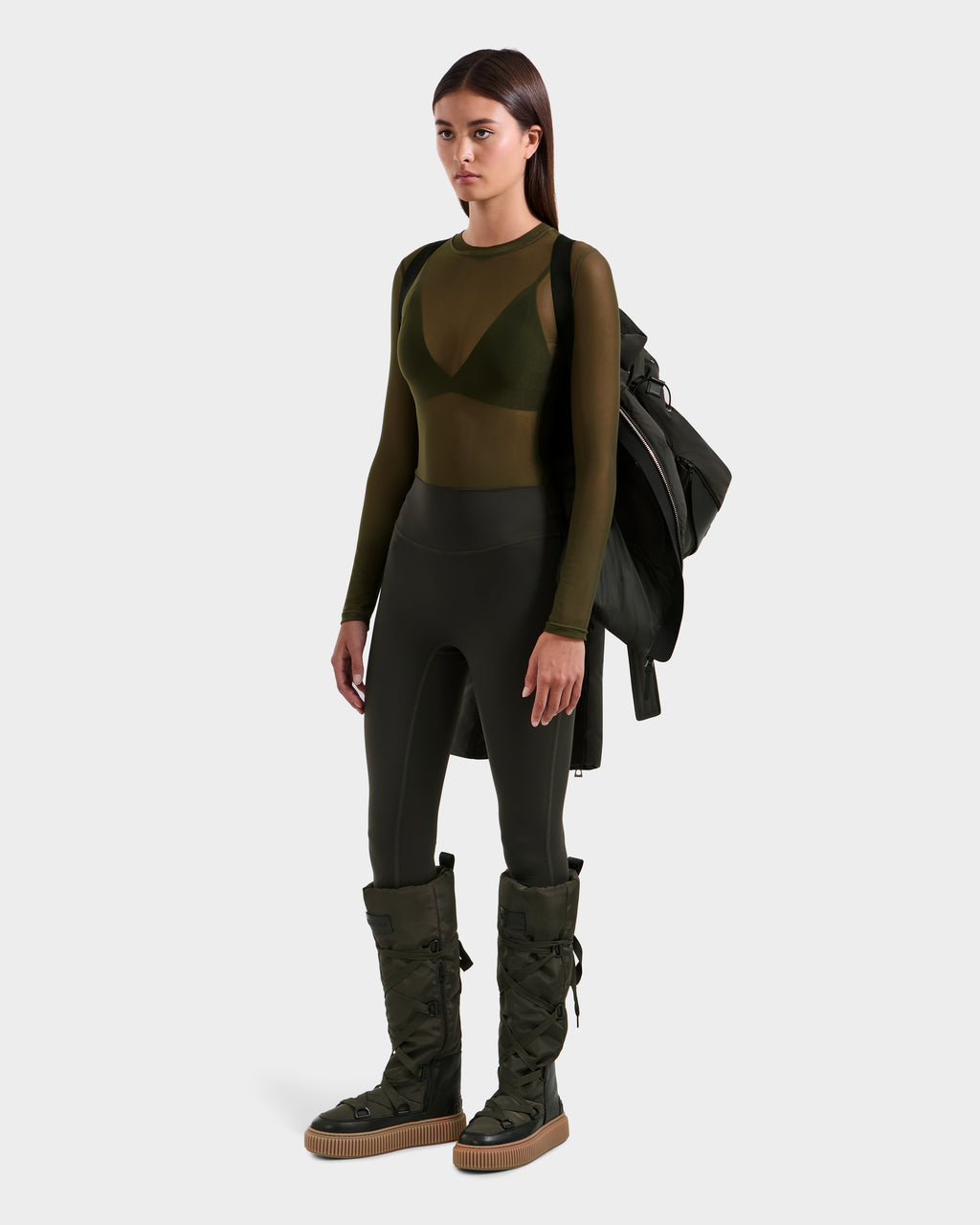 A woman faces forward, wearing an olive green sheer top with a bra, high-waisted leggings, lace-up boots, and carrying the Rudsak ADELINE H - 8125587 Dark Olive women’s down puffer jacket over her shoulder. The background is plain light gray.