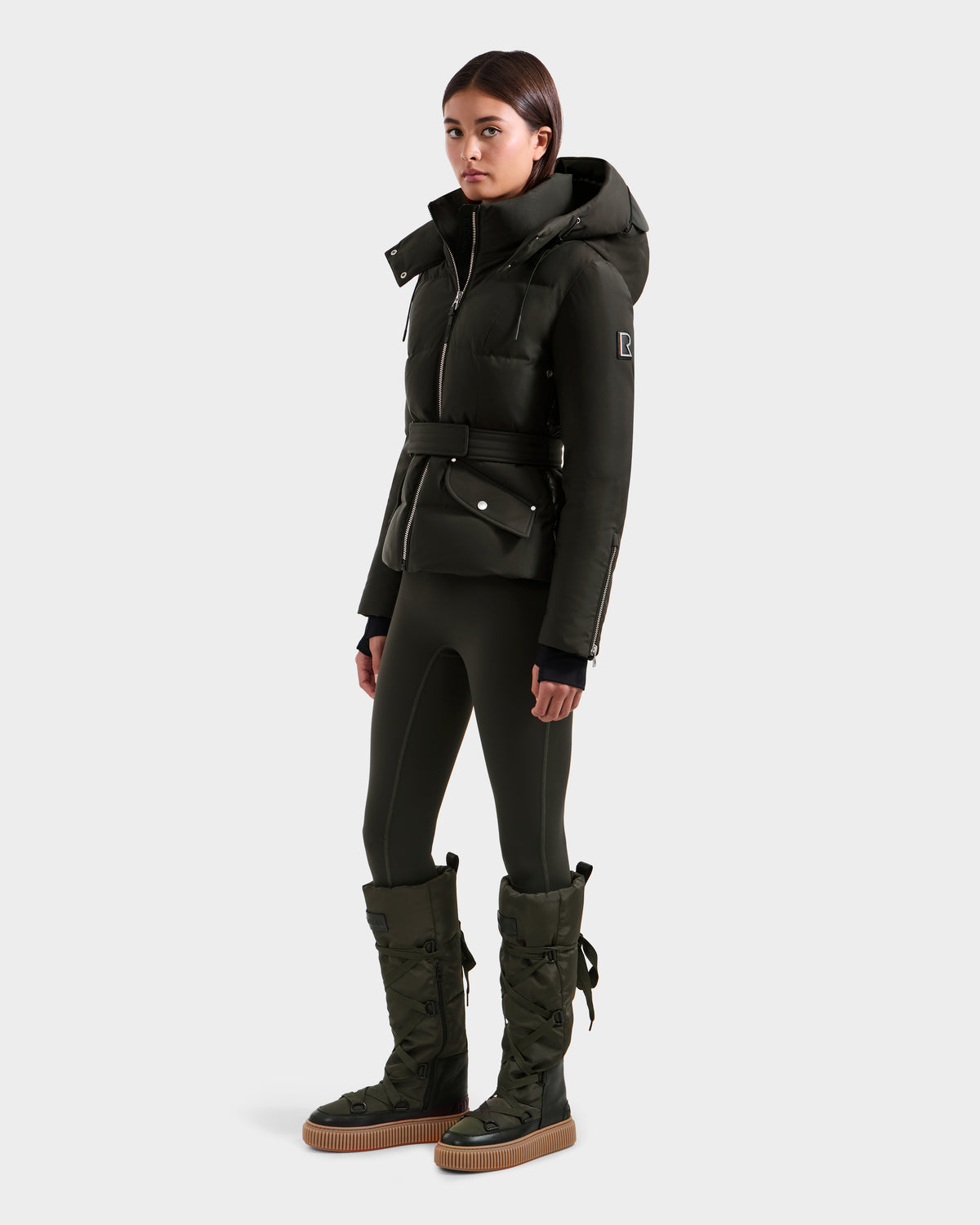 A woman stands against a light gray background wearing the Rudsak ADELINE H - 8125587 dark olive women’s down puffer jacket, matching leggings, and tall puffer winter boots.