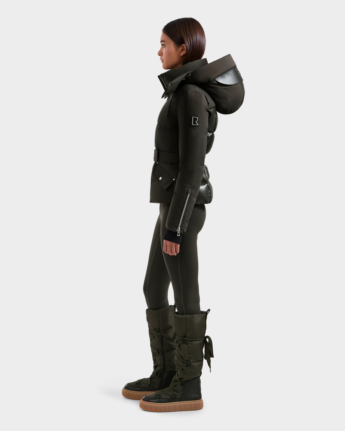 A person in profile wears the Rudsak ADELINE H - 8125587 women's dark olive down puffer jacket with a large hood, matching fitted pants, and tall quilted winter boots against a plain white background.