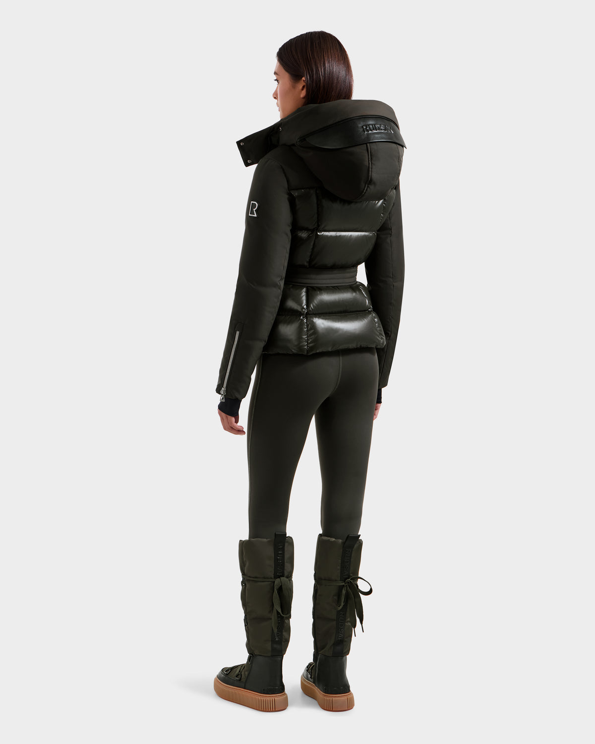 A woman faces away, wearing a shiny dark olive Rudsak ADELINE H - 8125587 winter jacket with a hood, paired with black leggings and tall padded boots. The background is plain and light-colored.