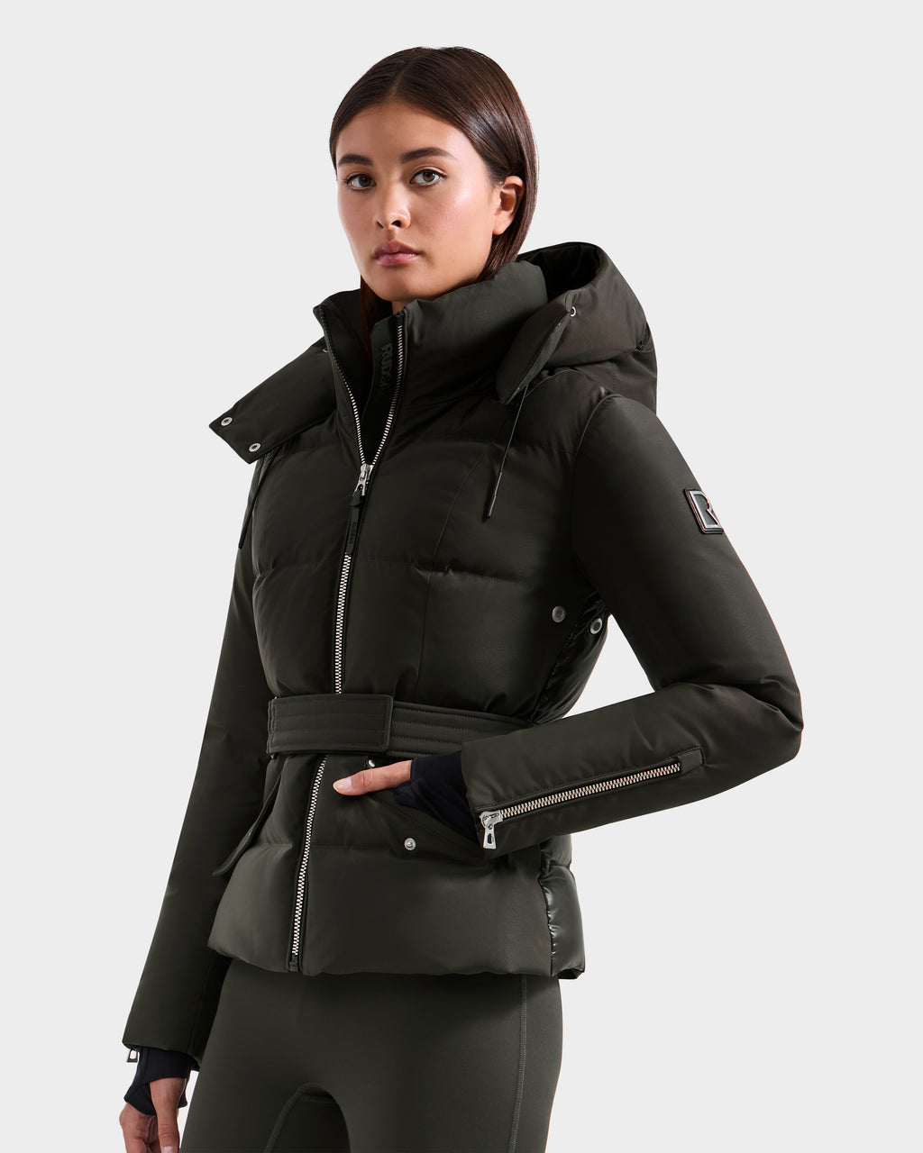 A woman in a Rudsak ADELINE H - 8125587 Dark Olive belted winter jacket with a hood and zippered sleeves stands against a plain light background, looking confidently at the camera.