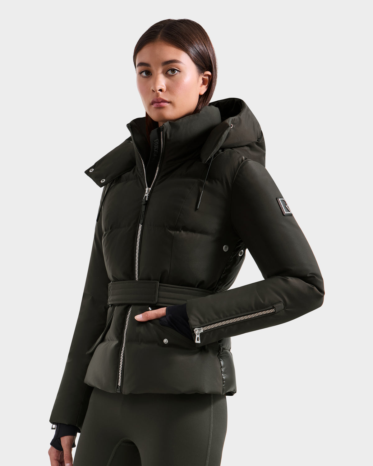 A woman in a Rudsak ADELINE H - 8125587 Dark Olive belted winter jacket with a hood and zippered sleeves stands against a plain light background, looking confidently at the camera.