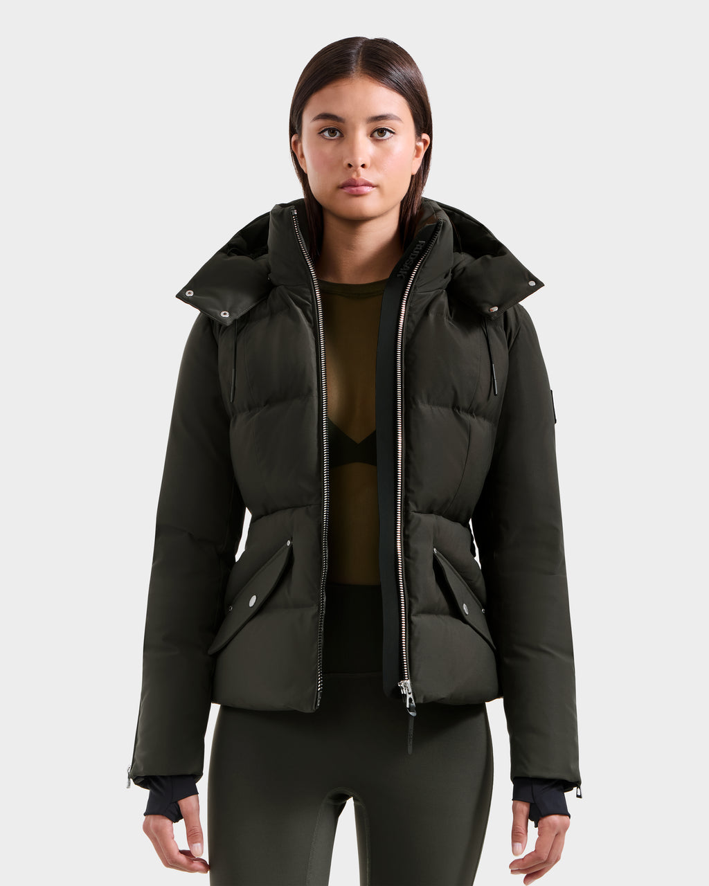 A woman with straight brown hair wears the Rudsak ADELINE H - 8125587 dark olive women's down puffer jacket over an olive top and leggings, standing against a plain, light gray background.
