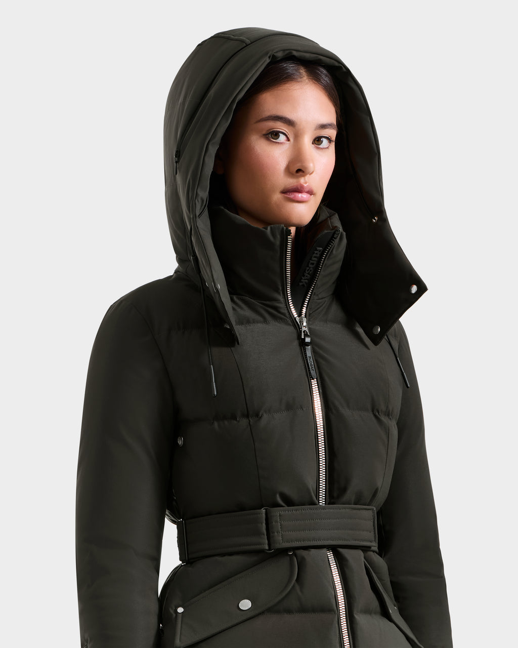 A woman wears a Rudsak ADELINE H - 8125587 dark olive women’s down puffer jacket with a large hood and belt. The zipped-up jacket stands out against a plain light gray background as she looks slightly to the side.