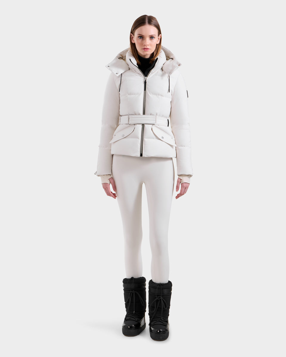 A woman faces forward against a white background, wearing the Rudsak ADELINE H - 8125587 Vanilla women's down puffer jacket with a belt, paired with white fitted pants and black winter boots.