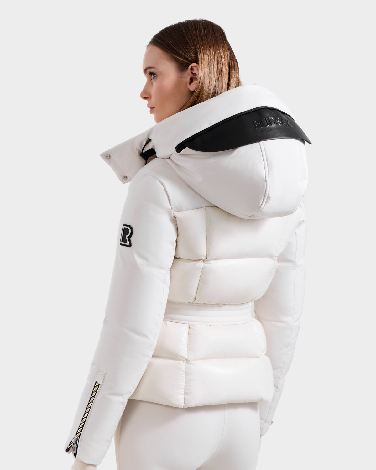 A woman with straight brown hair models the Rudsak ADELINE H - 8125587 Vanilla, a white women's down puffer with a large hood, black patch accents, and zippered sleeves, angled away against a light background.