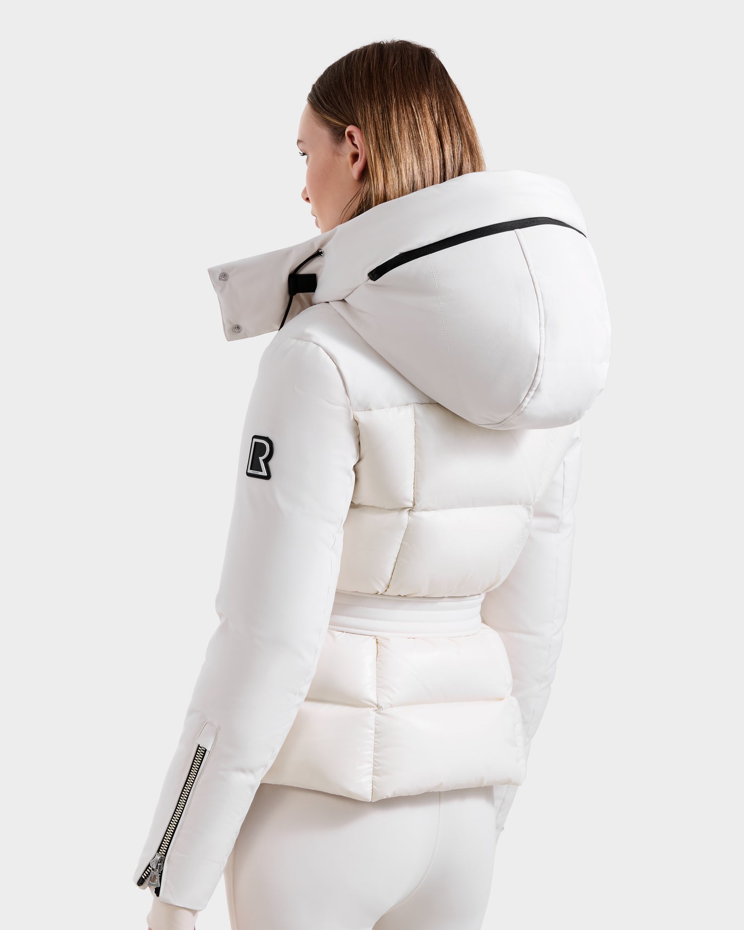 A person with straight, shoulder-length brown hair wears a Rudsak ADELINE H - 8125587 Vanilla jacket with a large hood, black sleeve logo patch, and zippered cuffs, photographed from the back against a plain background.