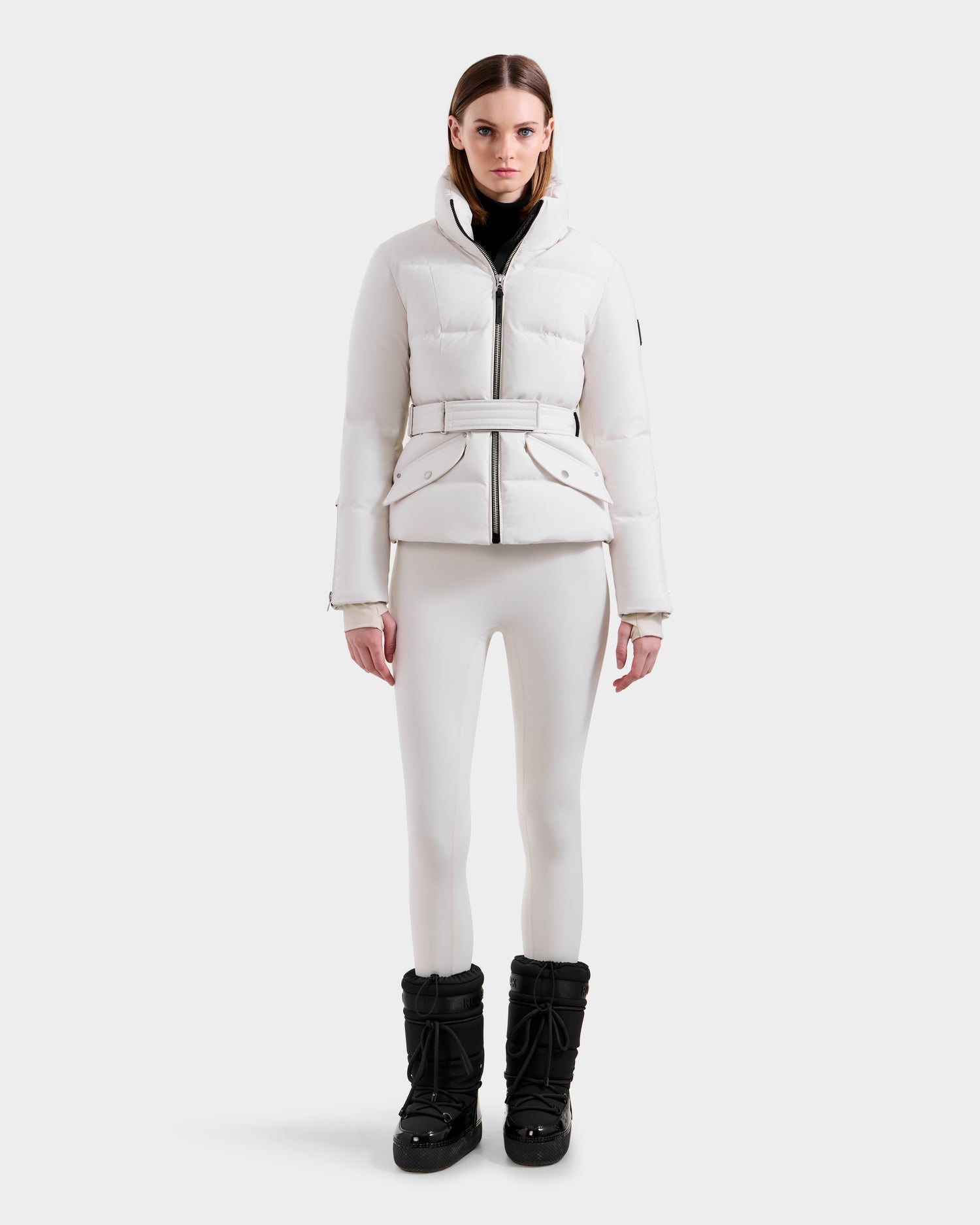 A woman faces forward in the Rudsak ADELINE H - 8125587 Vanilla fitted white down puffer coat with a belt, matching white leggings, black turtleneck, and black boots against a plain background, showcasing this stylish winter jacket.