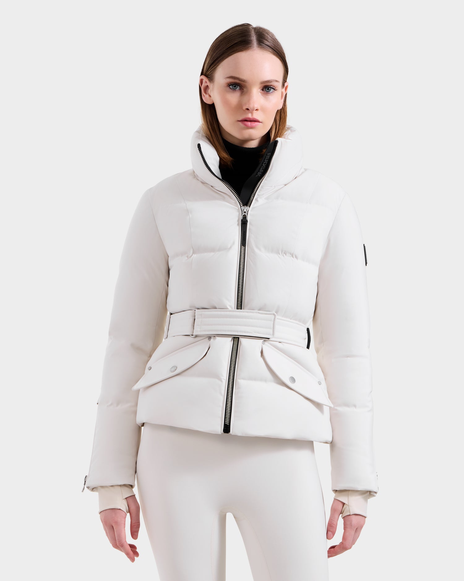 A woman wears the Rudsak ADELINE H - 8125587 Vanilla jacket with a high collar, black zipper, belted waist, and flap pockets, styled with white pants and a black turtleneck for an elegant down puffer look.