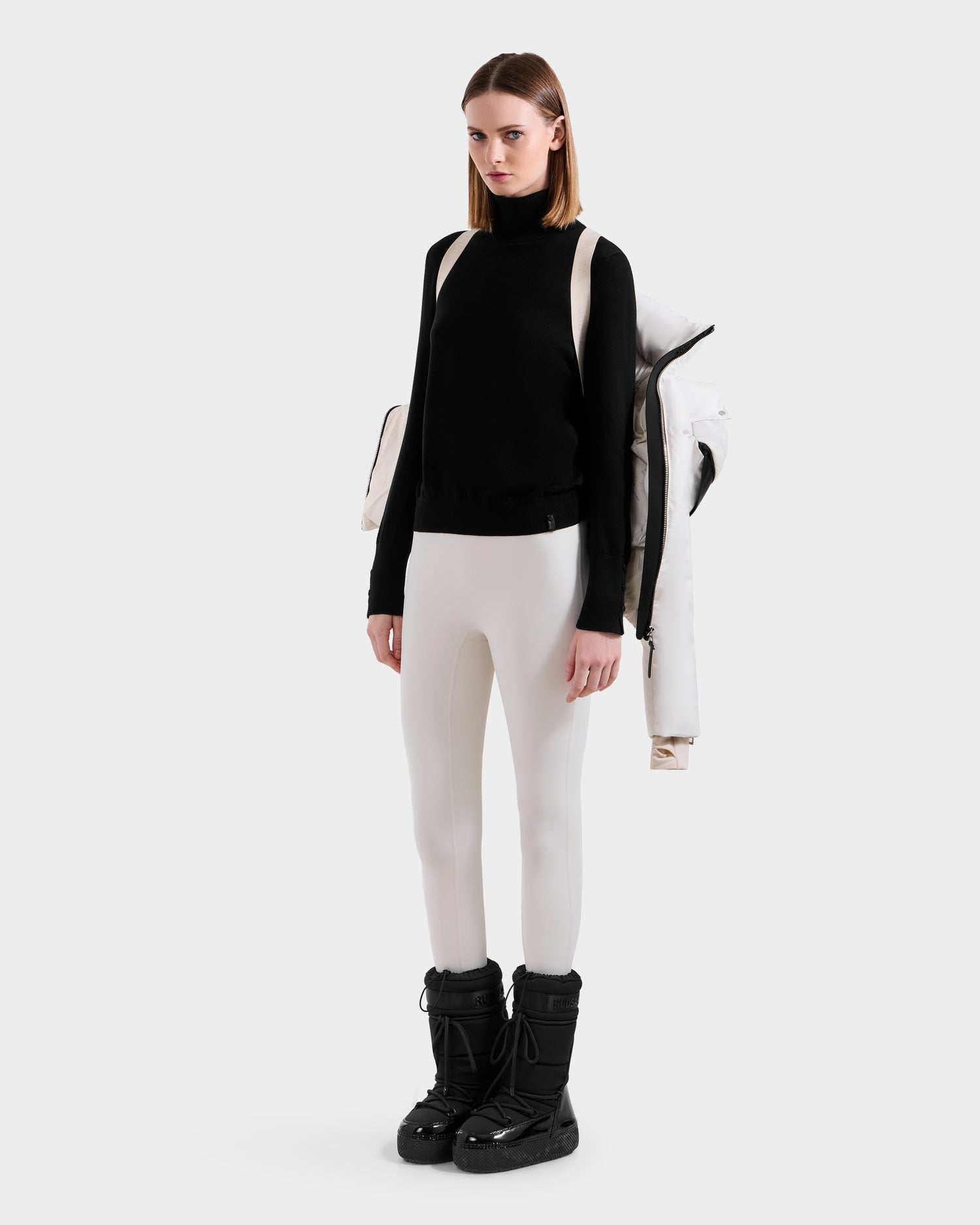 A woman stands against a plain background wearing a black turtleneck, white fitted pants, black snow boots, and carries the Rudsak ADELINE H - 8125587 Vanilla women’s down puffer jacket draped over her shoulder using backpack-style straps.