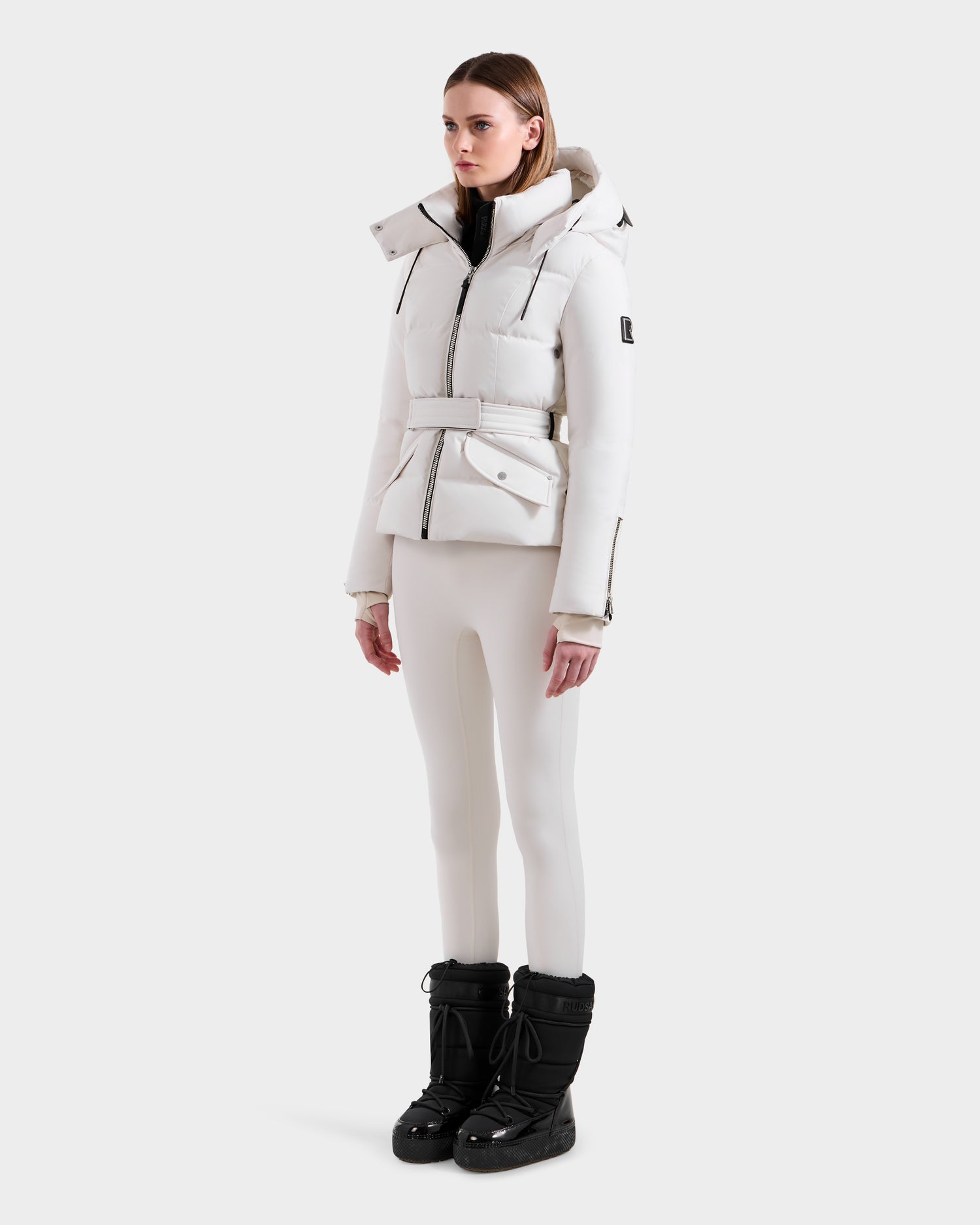 A woman with straight brown hair and a neutral expression stands against a plain background wearing the Rudsak ADELINE H - 8125587 Vanilla down puffer jacket, off-white pants, and black winter boots.
