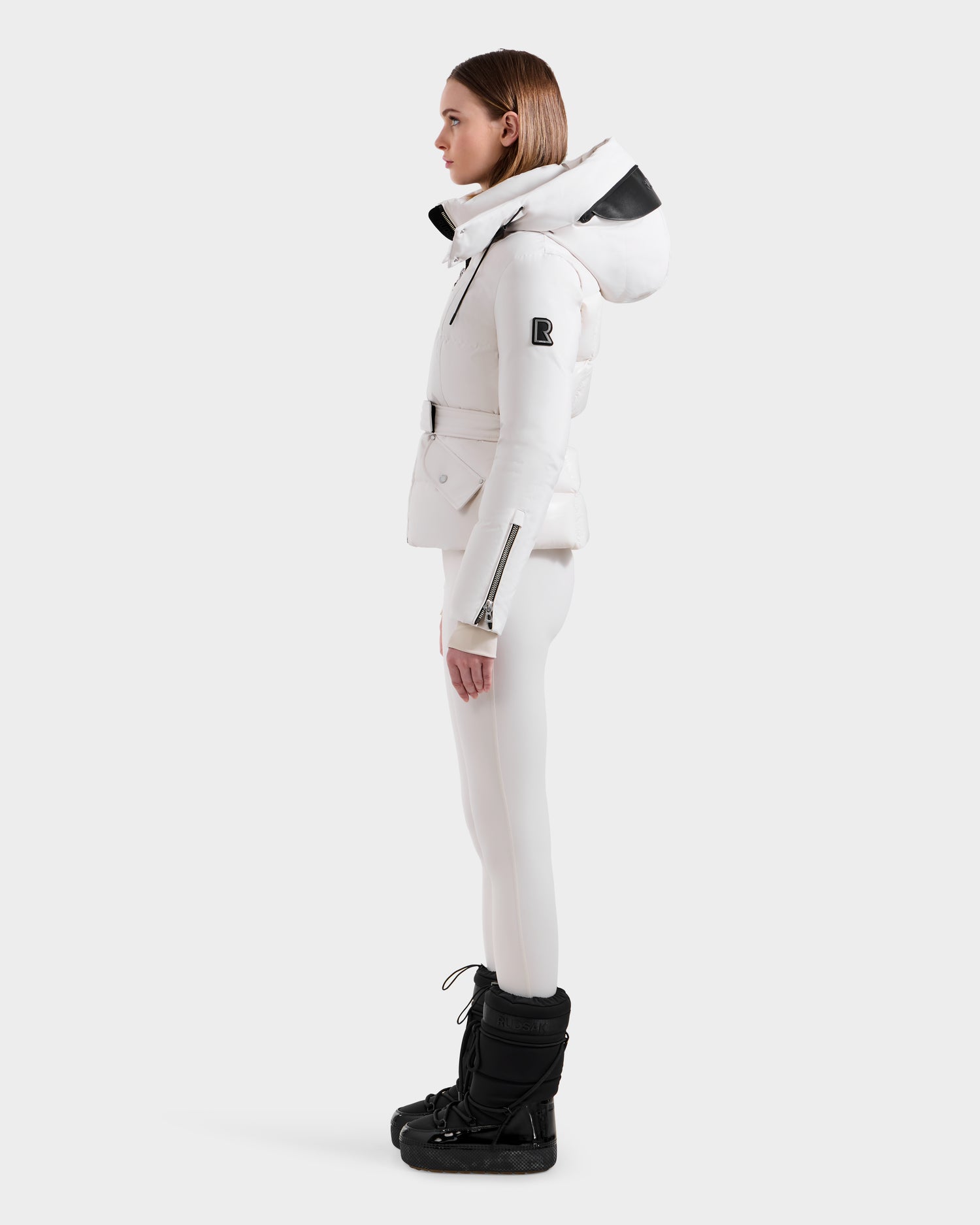 A person stands in profile wearing the Rudsak ADELINE H - 8125587 Vanilla women's down puffer jacket, paired with white pants and black winter boots against a plain white background.