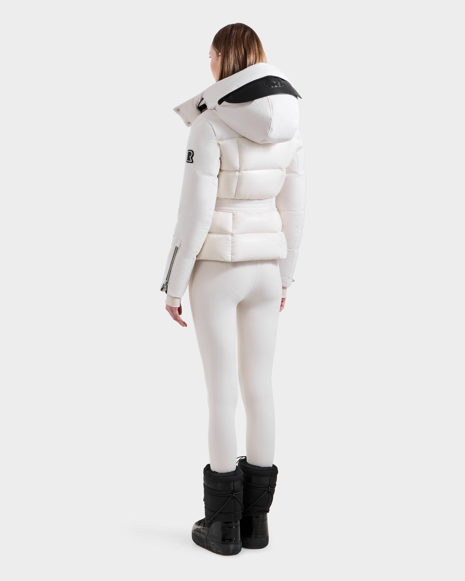 A woman faces away, wearing a vanilla Rudsak ADELINE H - 8125587 women’s down puffer jacket with a hood, paired with white leggings and black winter boots against a plain light background.