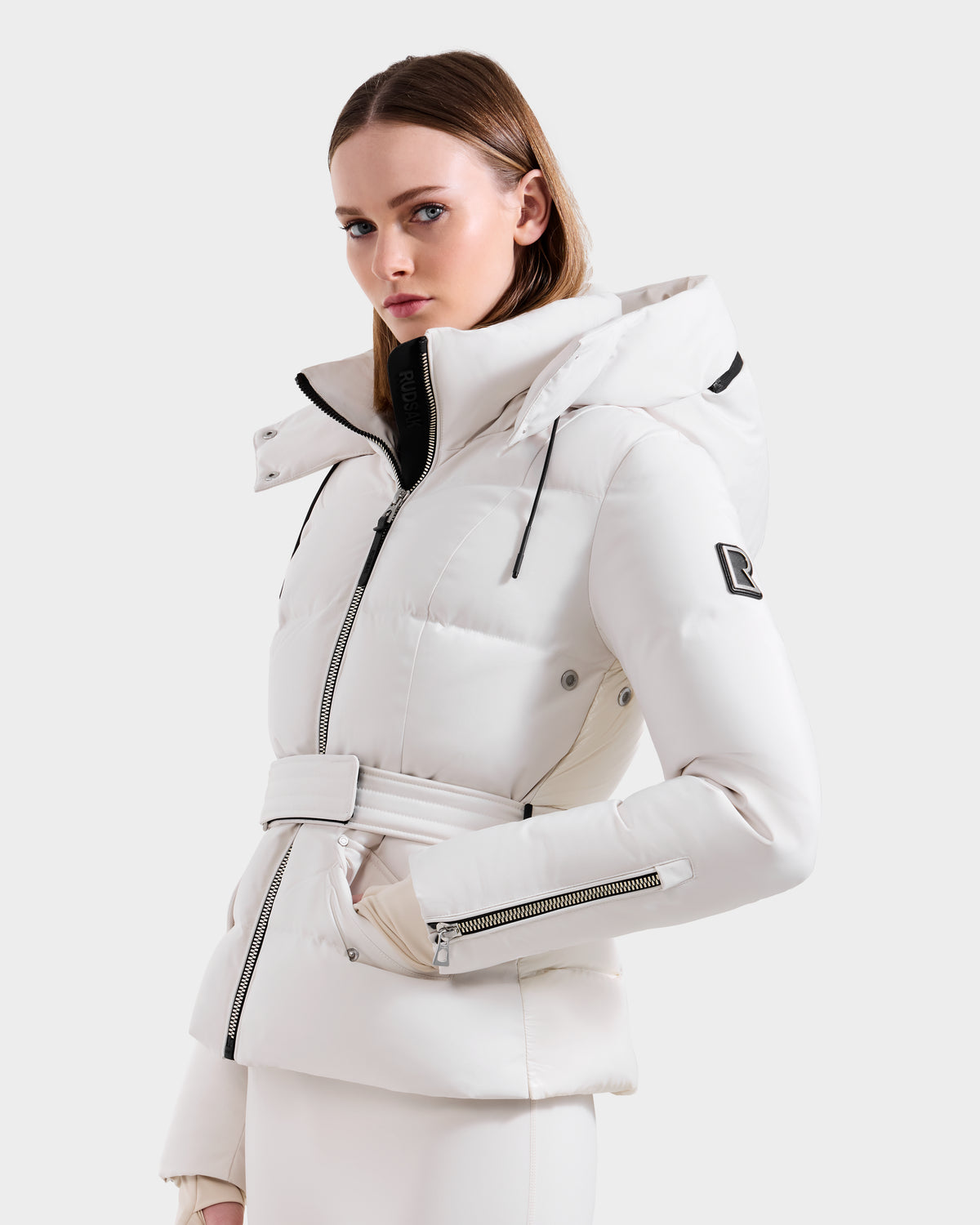 A woman models the ADELINE H - 8125587 Vanilla by Rudsak—a stylish white women’s down puffer with a belt, zippered pockets, high collar, hood, black logo patch on the sleeve, and silver zippers—against a plain white background.