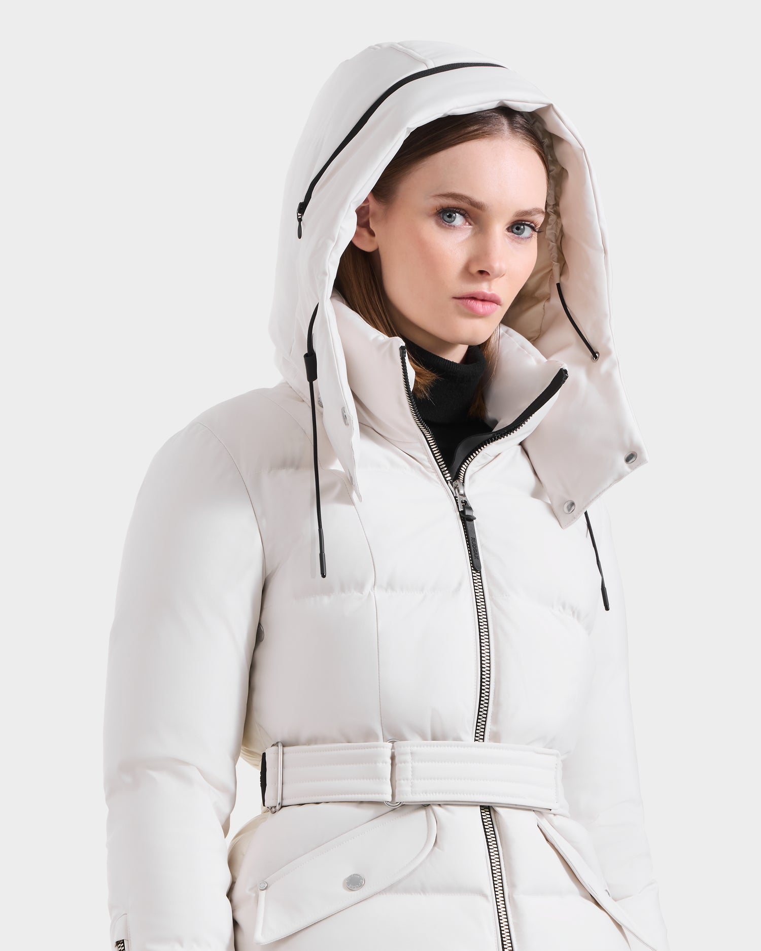 A woman wears the Rudsak ADELINE H - 8125587 Vanilla, a belted white down puffer with a large hood and black zipper accents, standing against a plain light background to showcase this stylish winter coat.