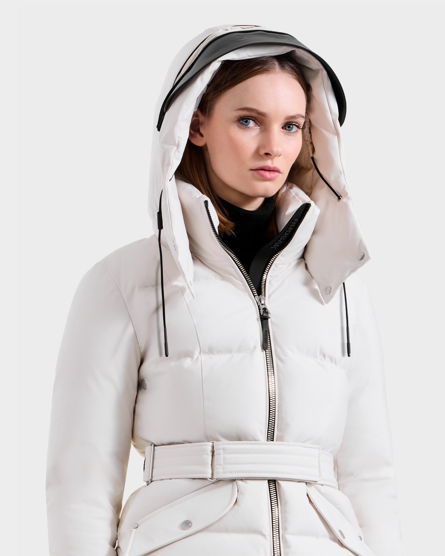 A woman wears the Rudsak ADELINE H - 8125587 Vanilla belted down puffer with hood and black zipper, looking slightly to the side against a plain light background.