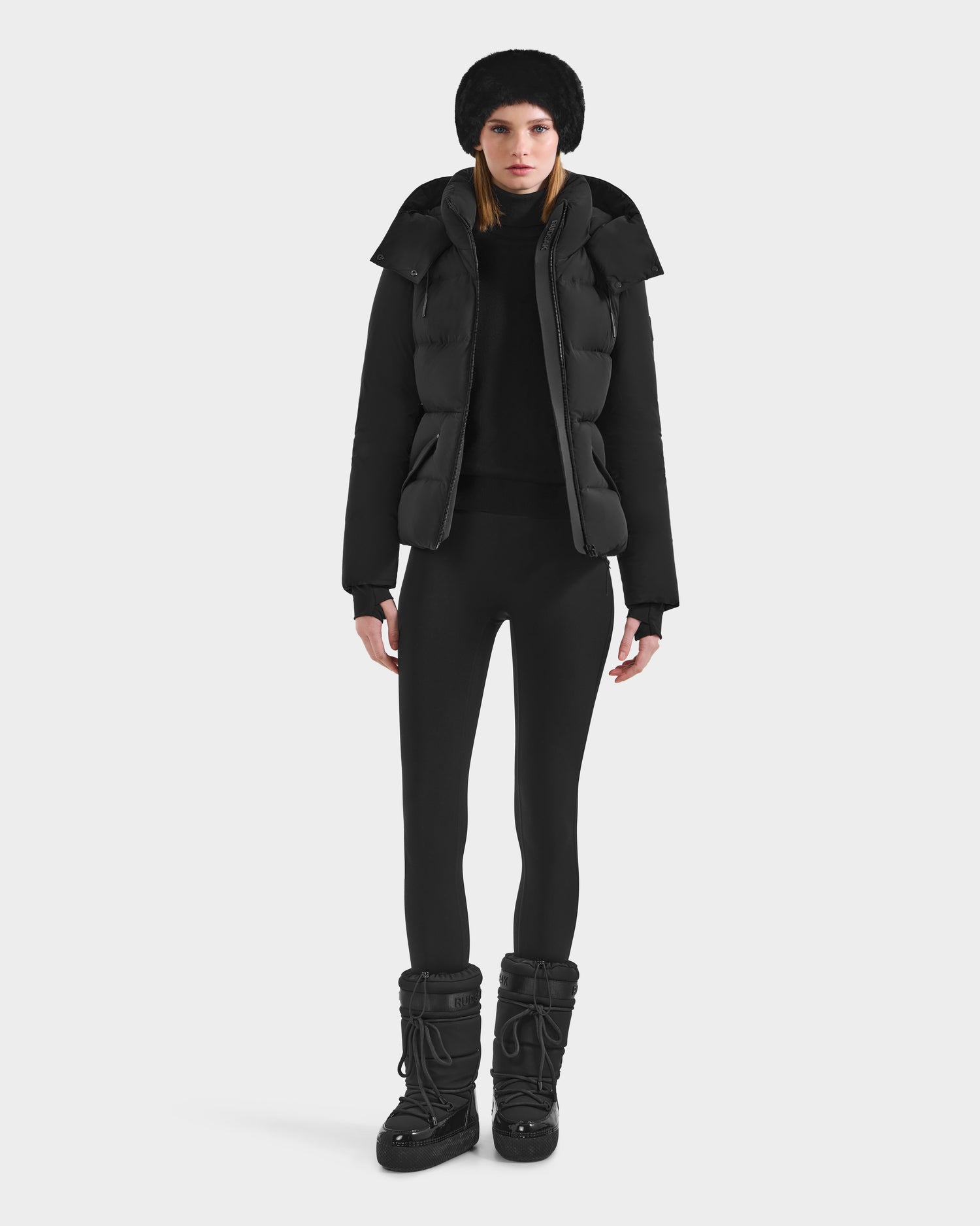 A woman stands facing forward, wearing the Rudsak ADELINE - 8124544 Black women's down puffer jacket, a black winter hat, leggings, and winter boots against a plain white background.