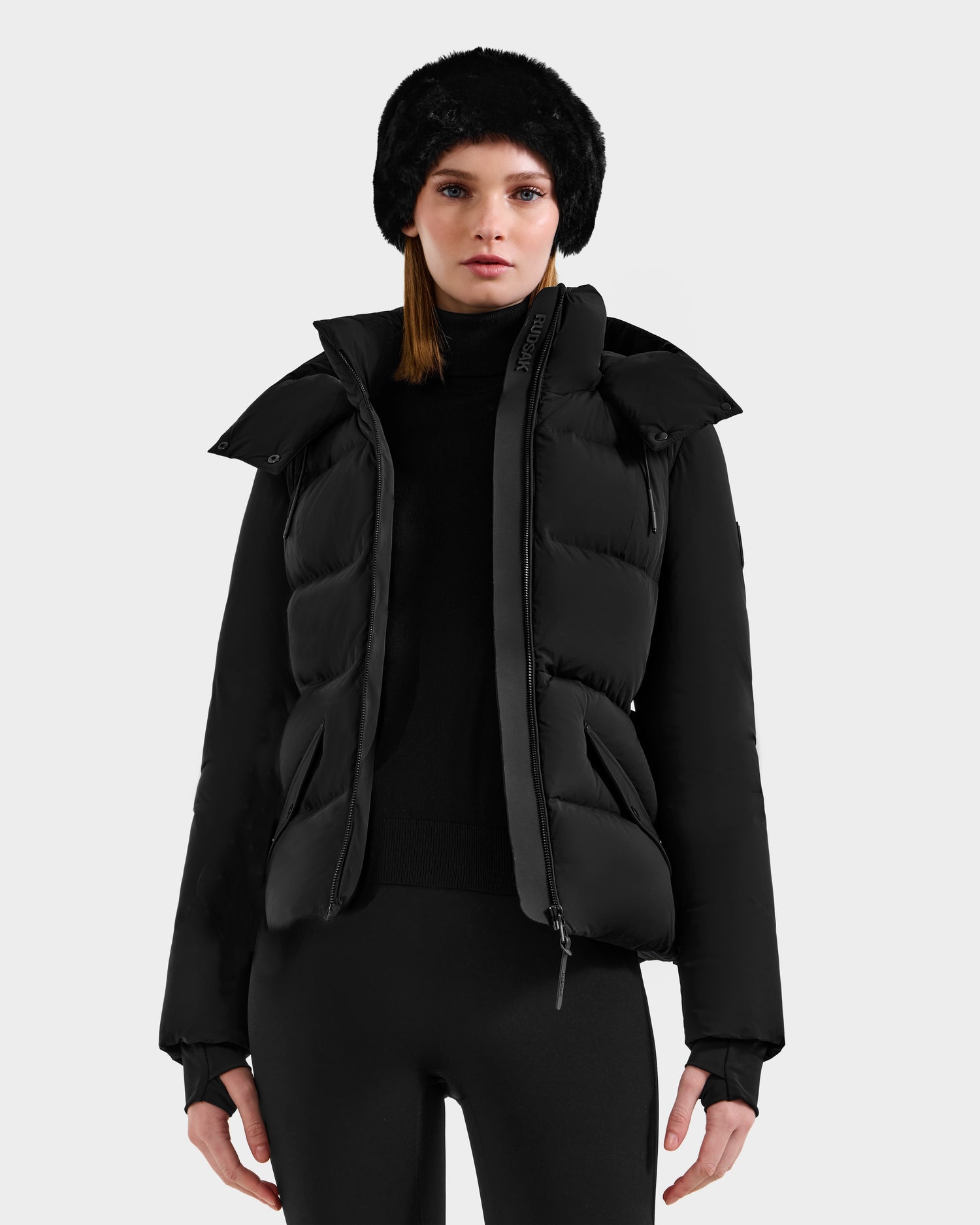 A woman wears the Rudsak ADELINE - 8124544 Black women's down puffer jacket, paired with a black turtleneck, leggings, and a black fur hat, standing against a plain light background.