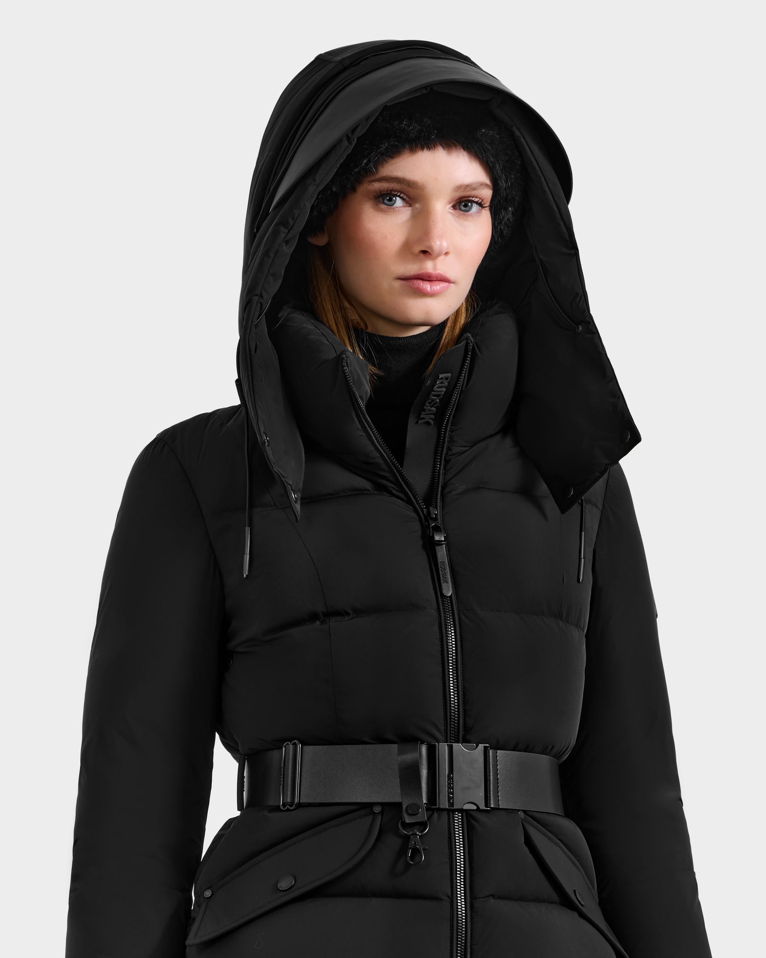 A woman stands against a plain light background, wearing the Rudsak ADELINE - 8124544 Black puffer jacket with an RDS-certified down fill, hood, black belt, and a black knit hat.