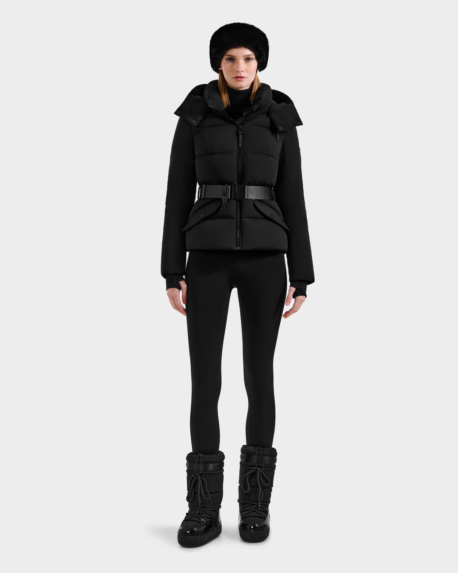 A woman faces forward wearing the Rudsak ADELINE - 8124544 Black down puffer with a belt, styled with black leggings, black boots, and a black fur hat, set against a plain light gray background.