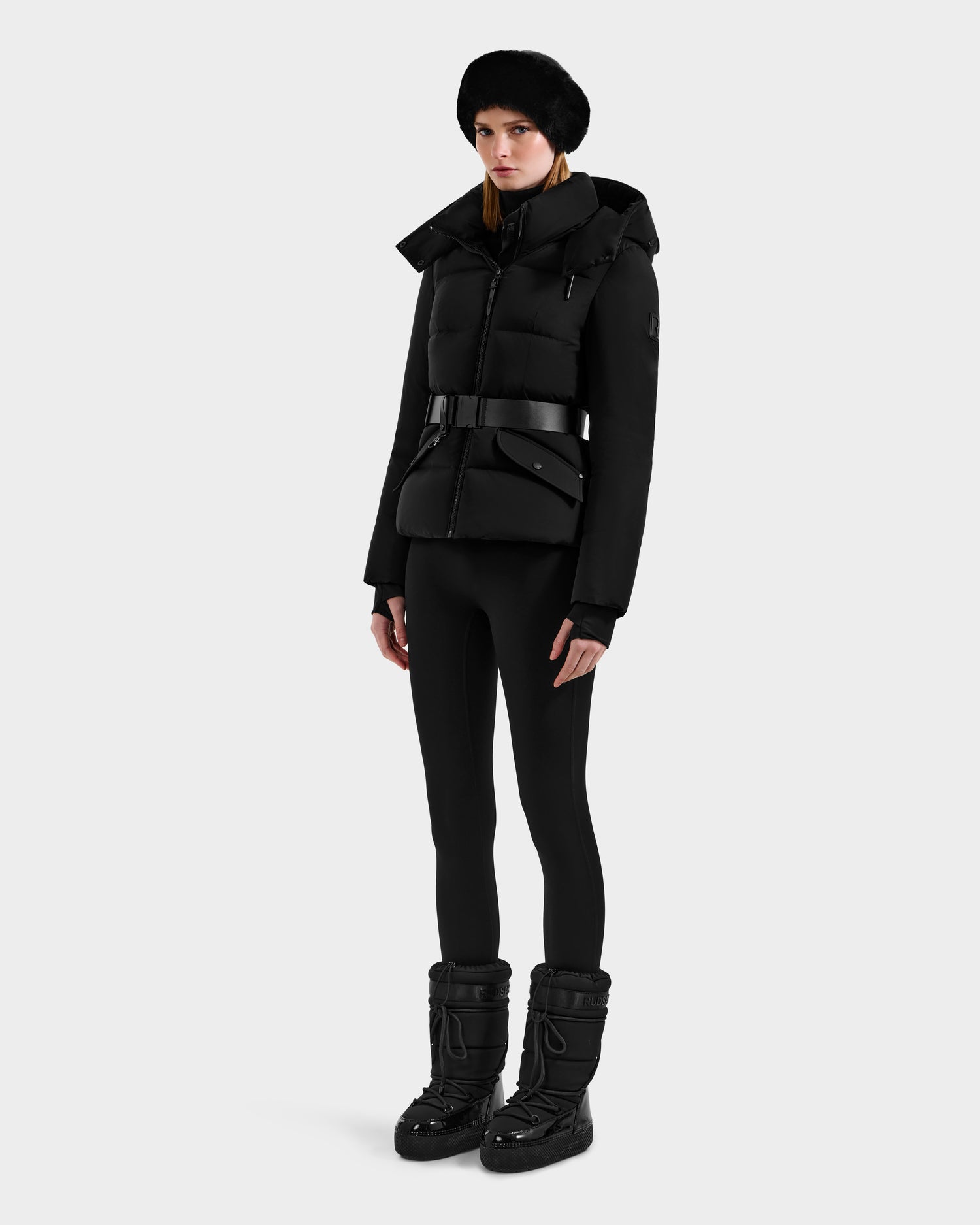 A woman models the Rudsak ADELINE - 8124544 Black down puffer jacket with a belt, paired with fitted leggings, snow boots, and a fur hat, against a light gray background.