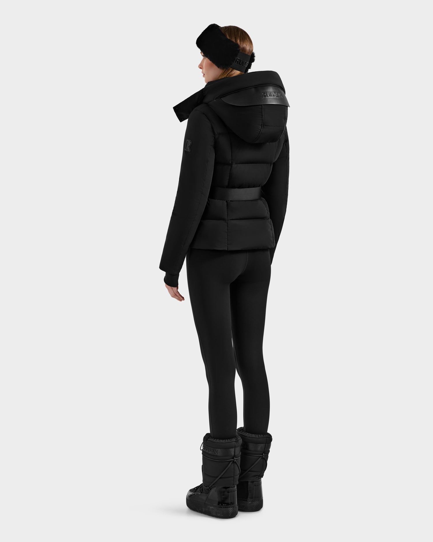 A person wearing the Rudsak ADELINE - 8124544 Black down puffer stands facing away in black leggings, snow boots, and a black headband against a plain white background.