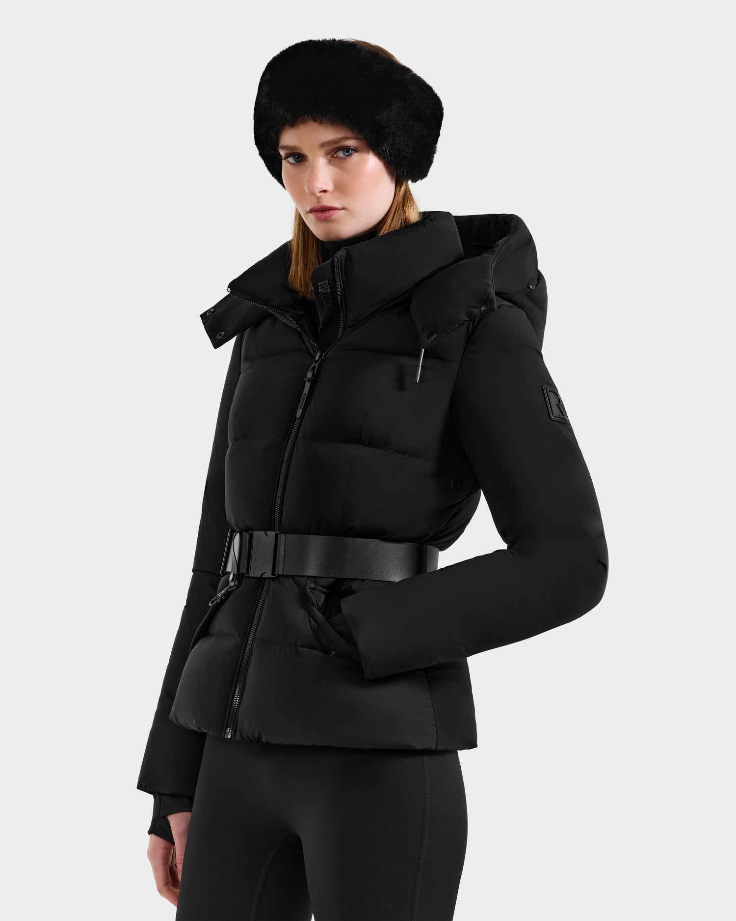A woman wearing the Rudsak ADELINE - 8124544 Black RDS-certified down puffer jacket with a wide belt, black leggings, and a black fur headband stands against a light gray background, gazing slightly to the side.