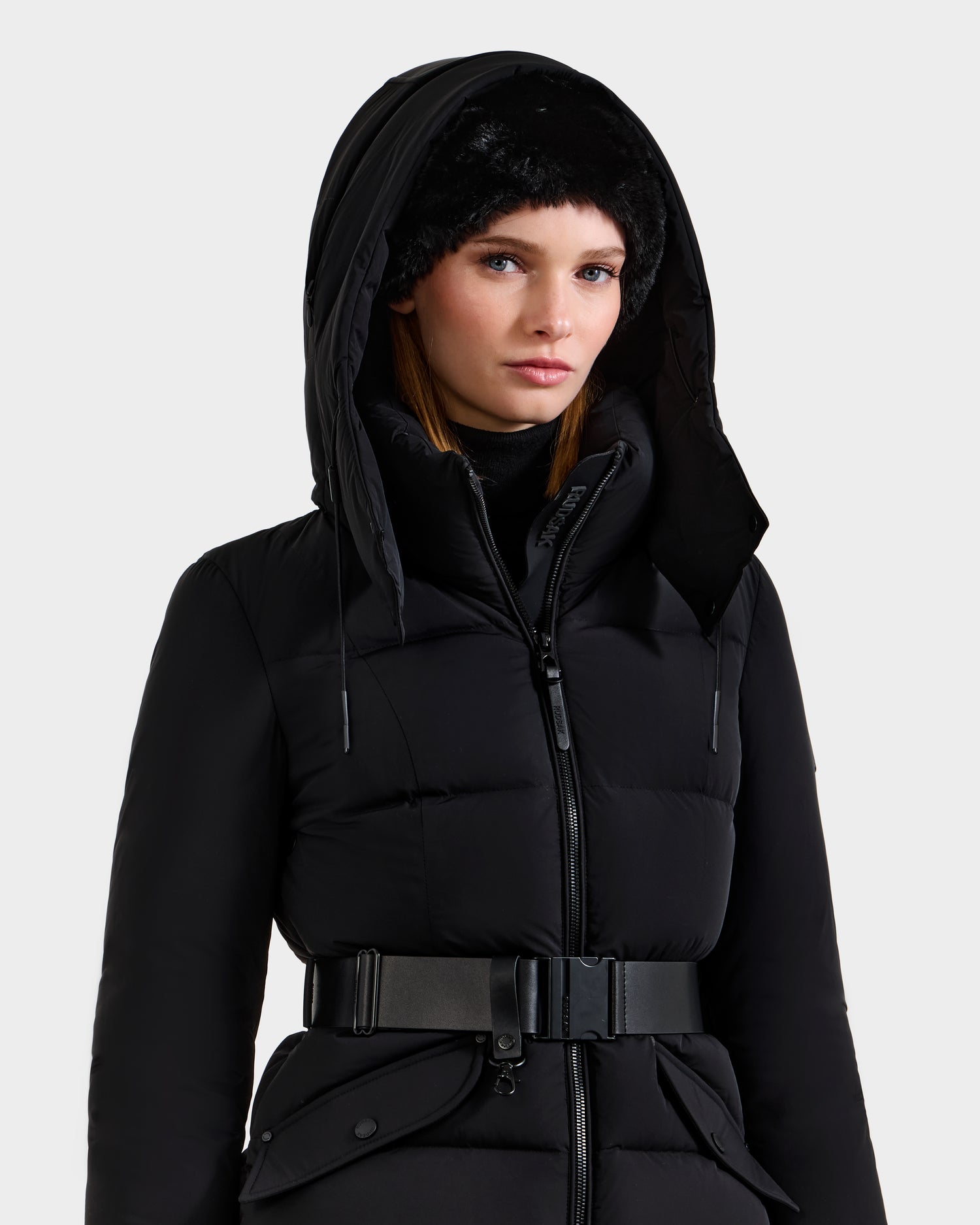 A woman wearing the Rudsak ADELINE - 8124544 Black quilted winter jacket with a wide belt and a black furry hat stands against a plain light background.