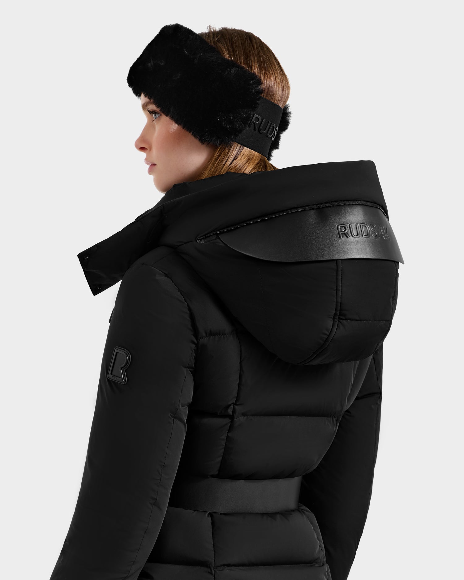 A woman is shown from the side and back wearing the Rudsak ADELINE - 8124544 Black women's down puffer jacket with a hood and a black fur headband, both featuring the Rudsak logo, against a plain light background.