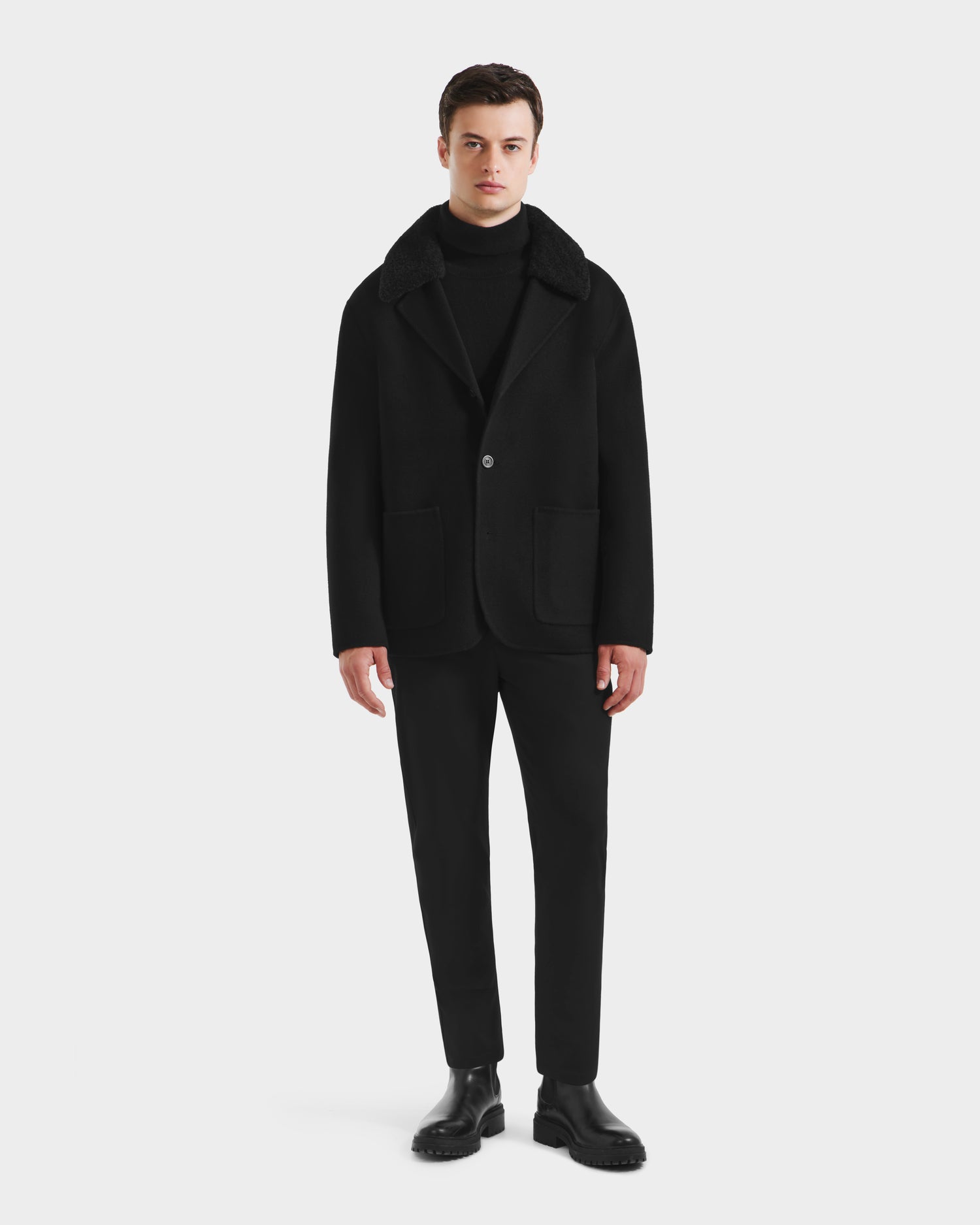 A man faces forward wearing the Rudsak AIDEN - 6125558 Black mens wool-blend coat with removable faux shearling collar, paired with a black turtleneck, trousers, and shoes against a plain white background.